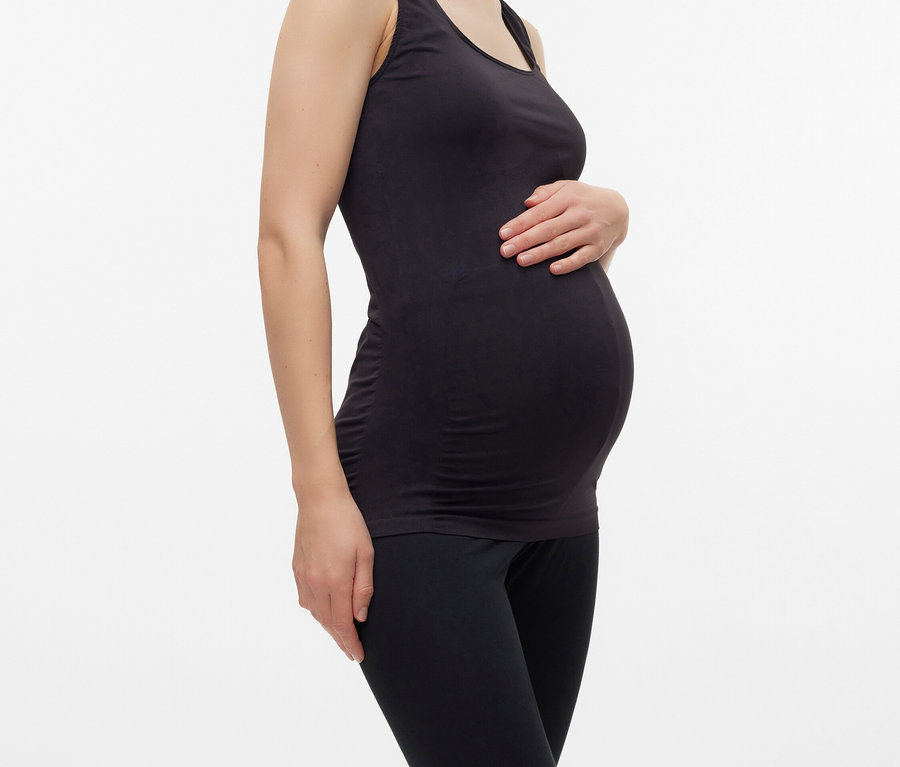 Tehotná žena v čiernom tielku a legínach pózuje na bielom pozadí.