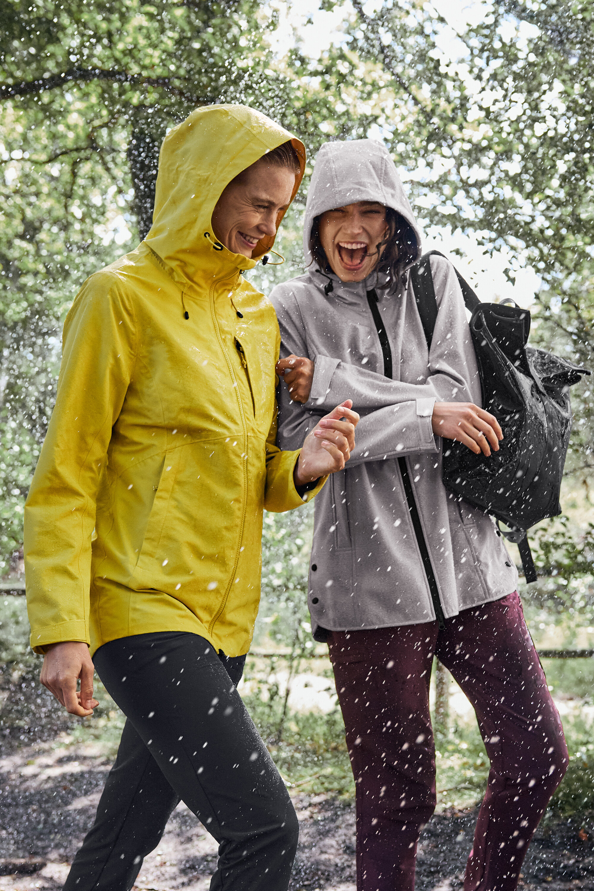 Dve ženy kráčajú v daždi, jedna má na sebe žltú pršiplášť, druhá sivú s čiernym batohom.