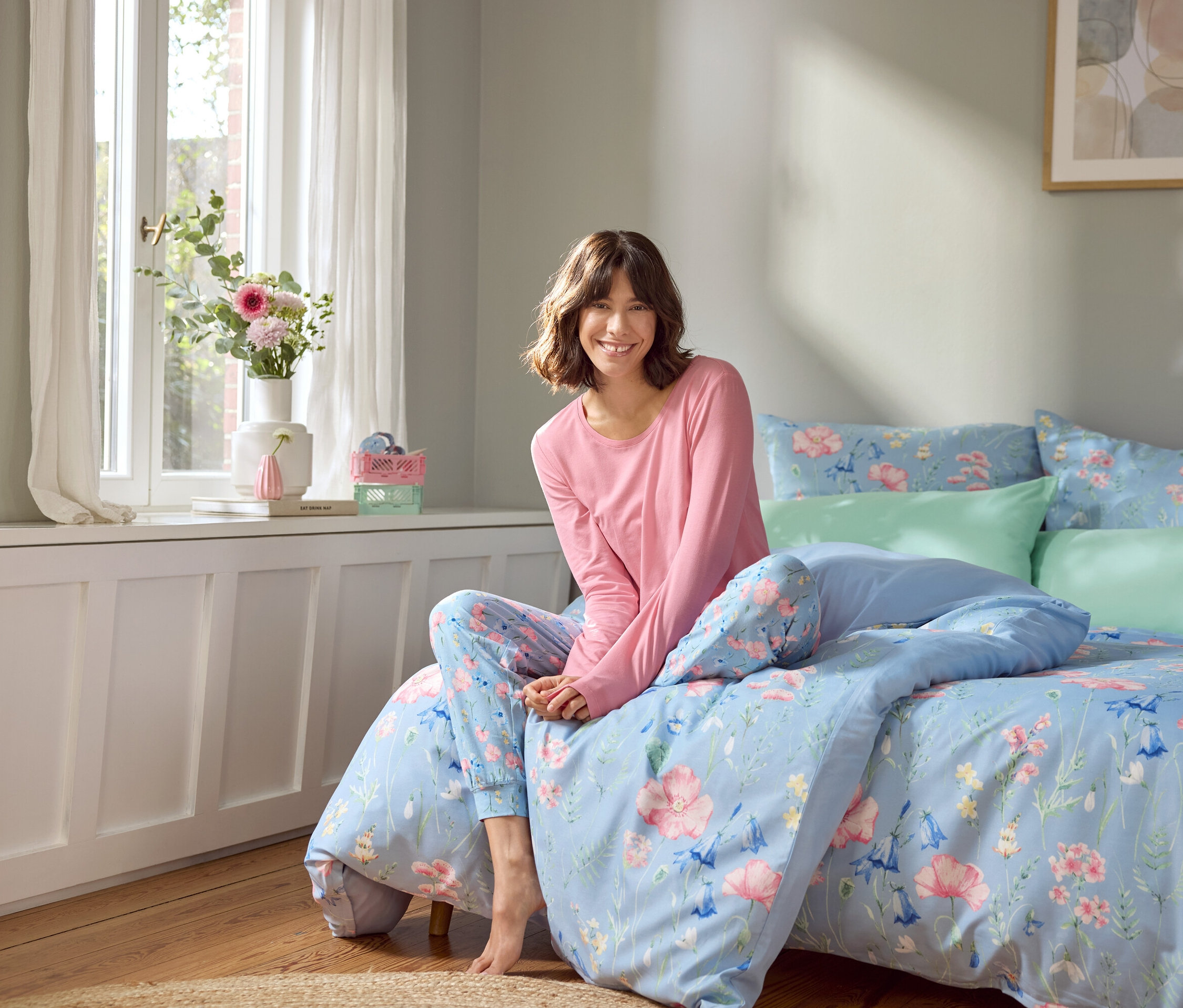 Žena sedí na posteli so svetlomodrou posteľnou bielizňou s kvetinovým vzorom.