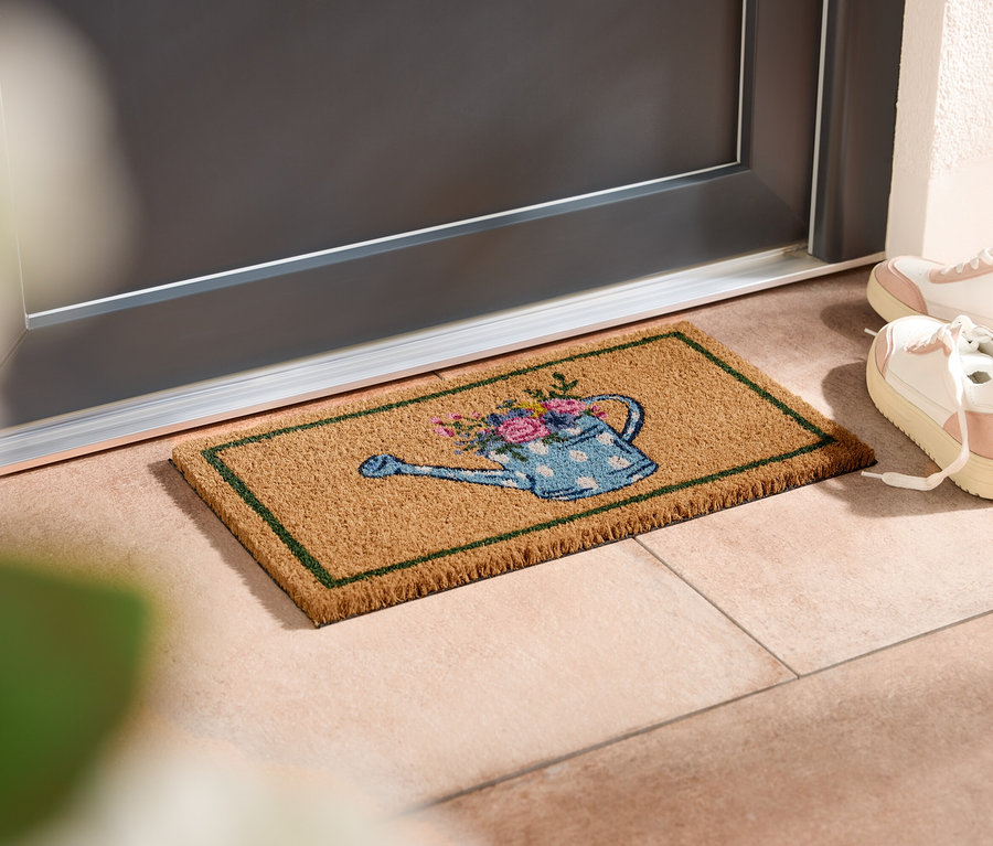 Rohožka s motívom kanvy na polievanie leží pred dverami, vedľa stoja biele tenisky.