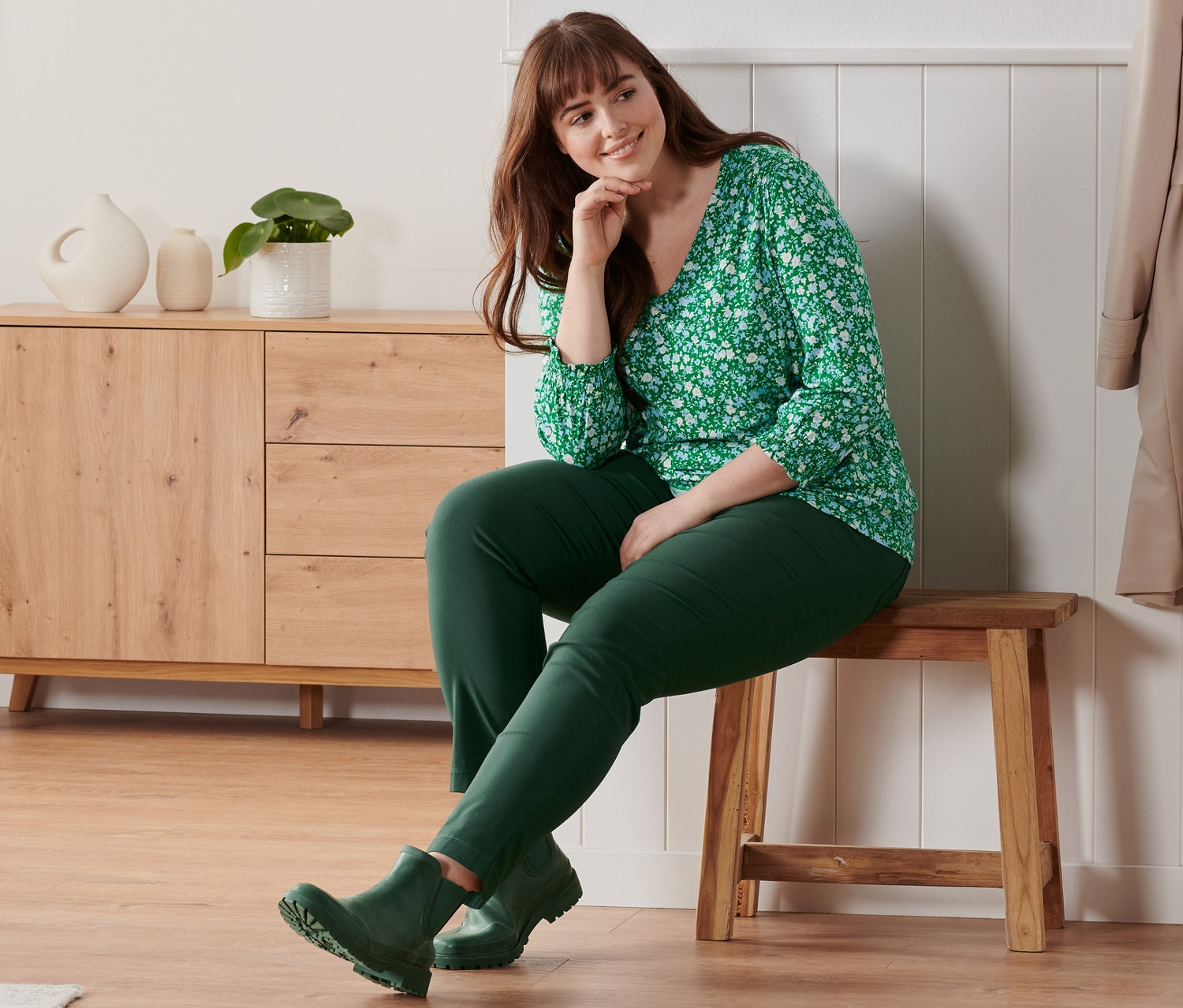 Žena sedí na lavici a má na sebe zelené blúzkové tričko s trojštvrťovým rukávom s celoplošnou kvetinovou potlačou, bengalínové nohavice a krátke čižmy do dažďa.