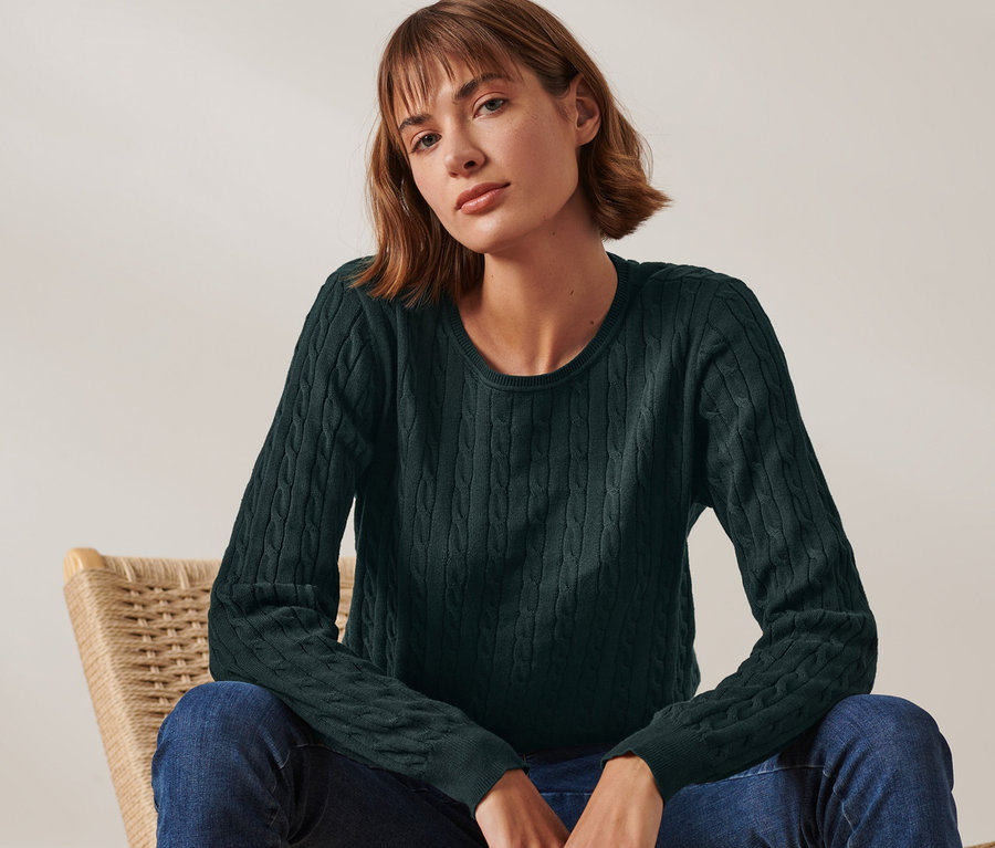 Žena sedí na prútenom kresle a má na sebe tmavozelený pletený pulóver s vrkočovým vzorom.