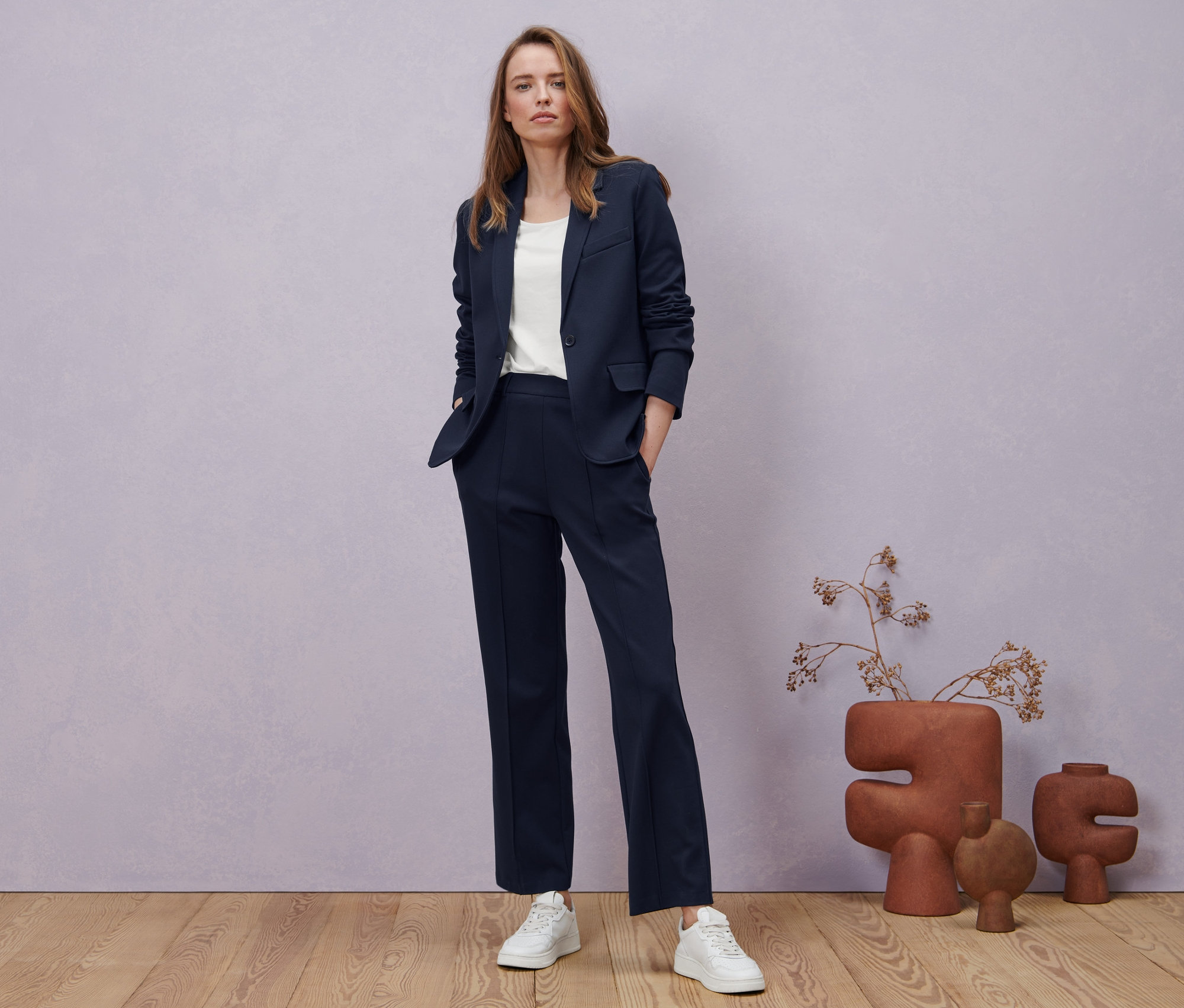 Žena pózuje v tmavomodrých džersejových nohaviciach so zámikom, džersejovom saku a bielom tričku s polodlhým rukávom.