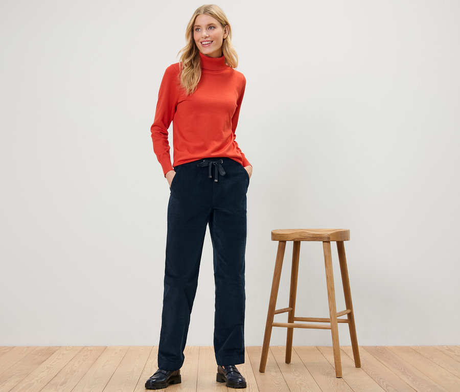 Žena stojí a pózuje v menčestrových pohodlných nohaviciach a oranžovom roláku.