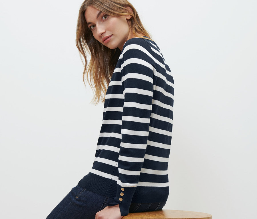 Žena sedí na drevenej stoličke v pruhovanom pulóvri z jemného úpletu, námornícka modrá a v džínsoch.