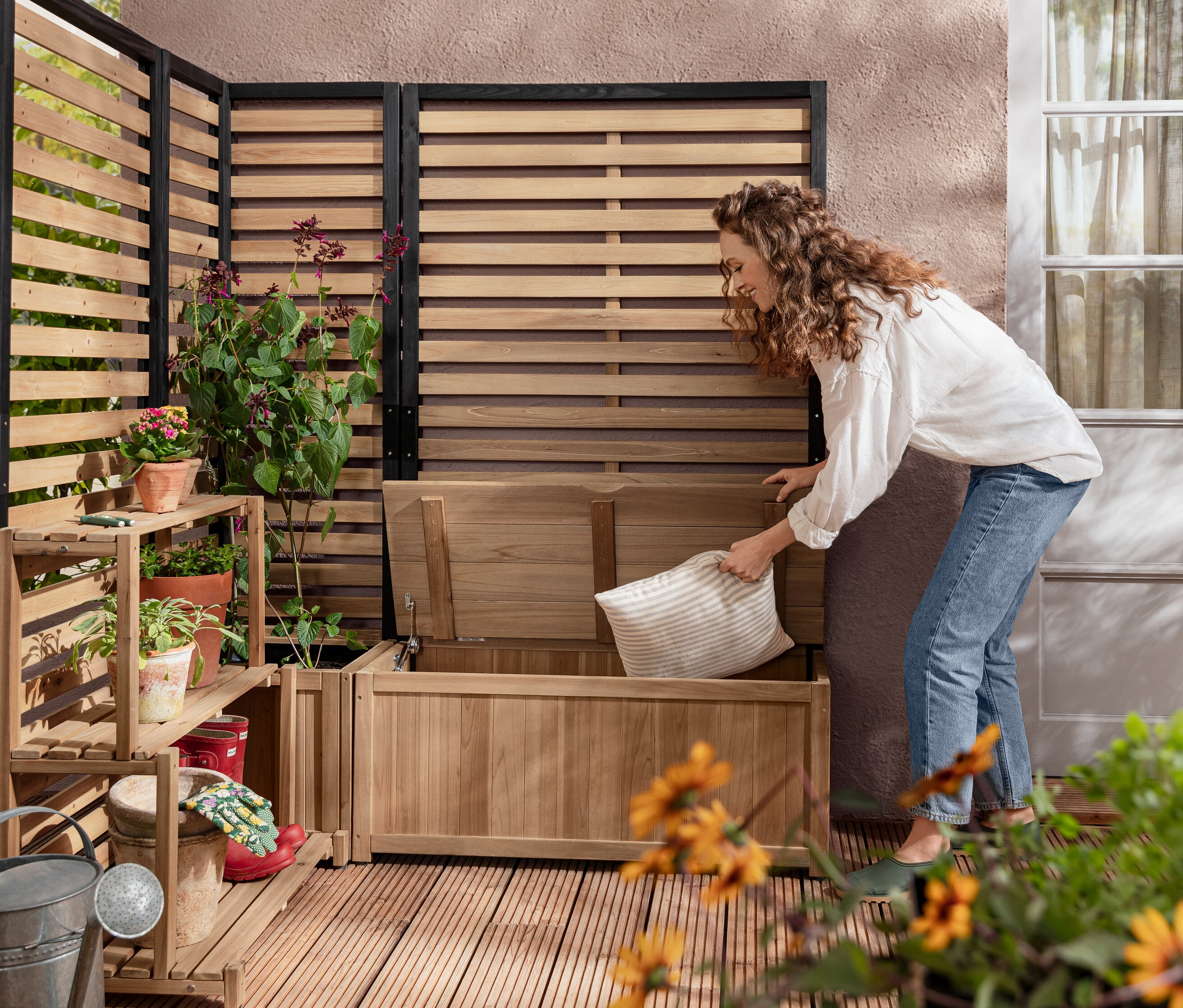 Žena vkladá vankúš do úložnej lavice Zástena s modulom odkladacej truhlice »Elin«. V pozadí je Modul »Elin«, zástena s kvetináčom a Zástenový regálový modul »Elin«.