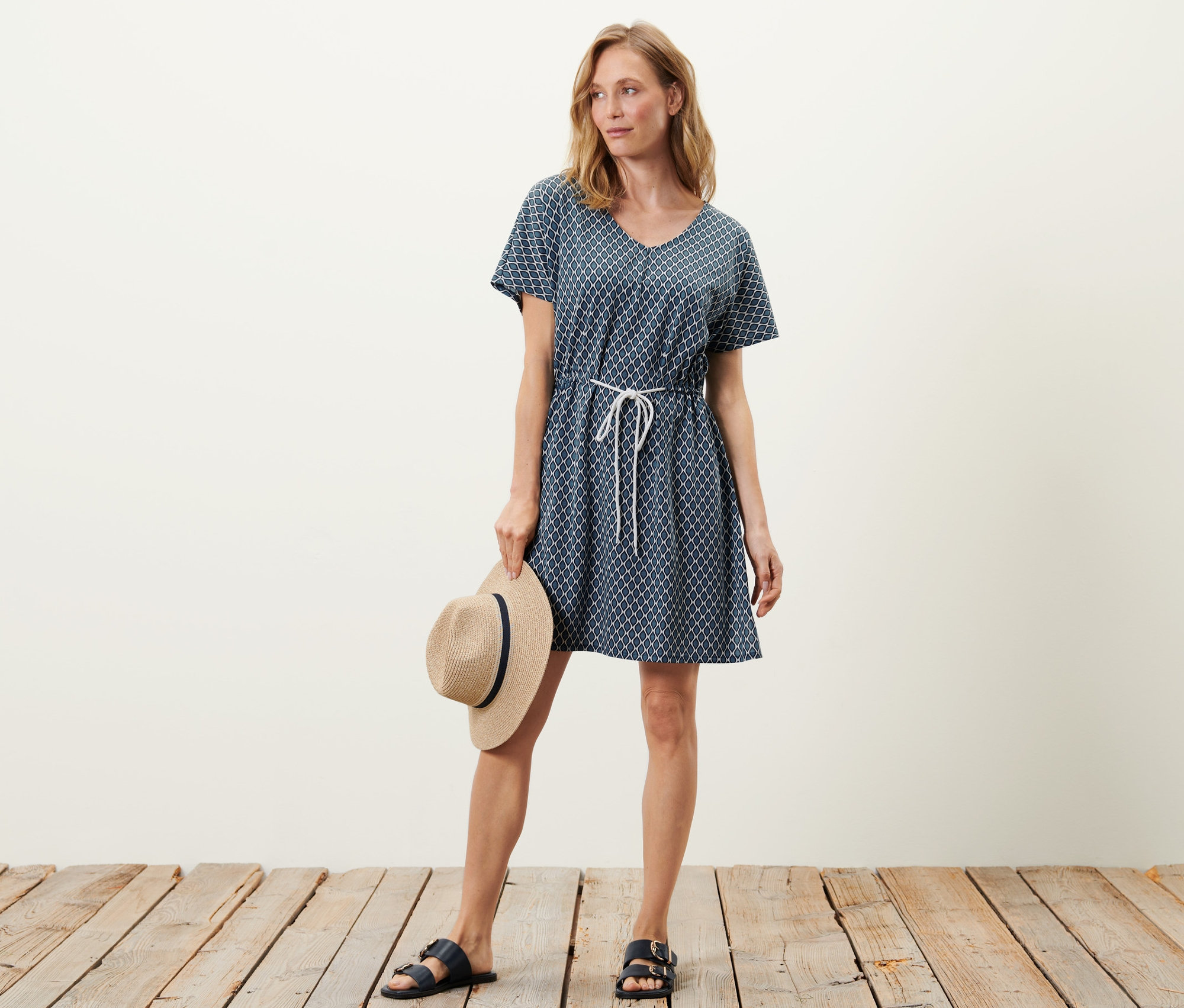 Žena pózuje v plážovej tunike a drží klobúk.