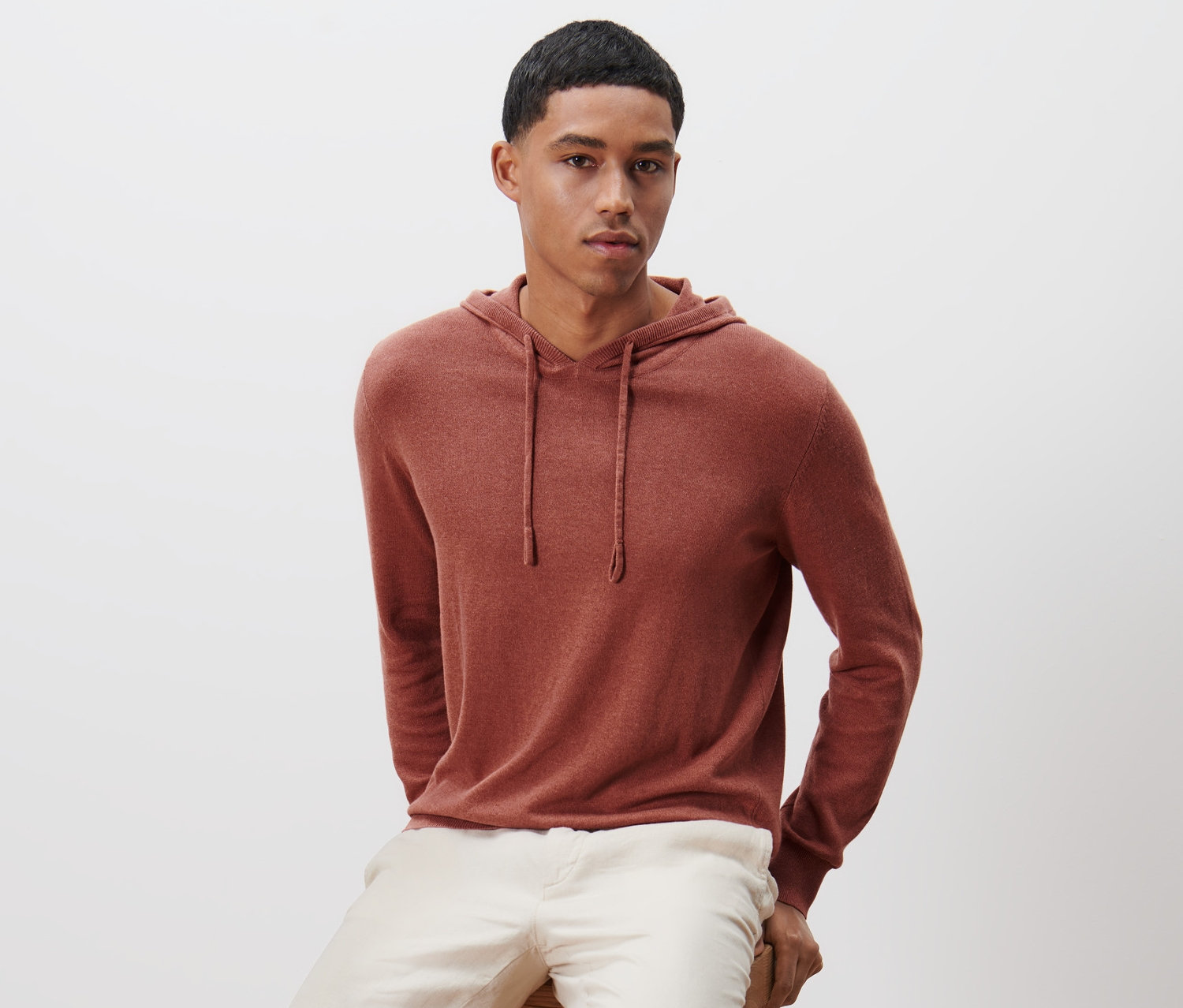 Muž sedí na drevenej stoličke a má na sebe pletenú mikinu s kapucňou.