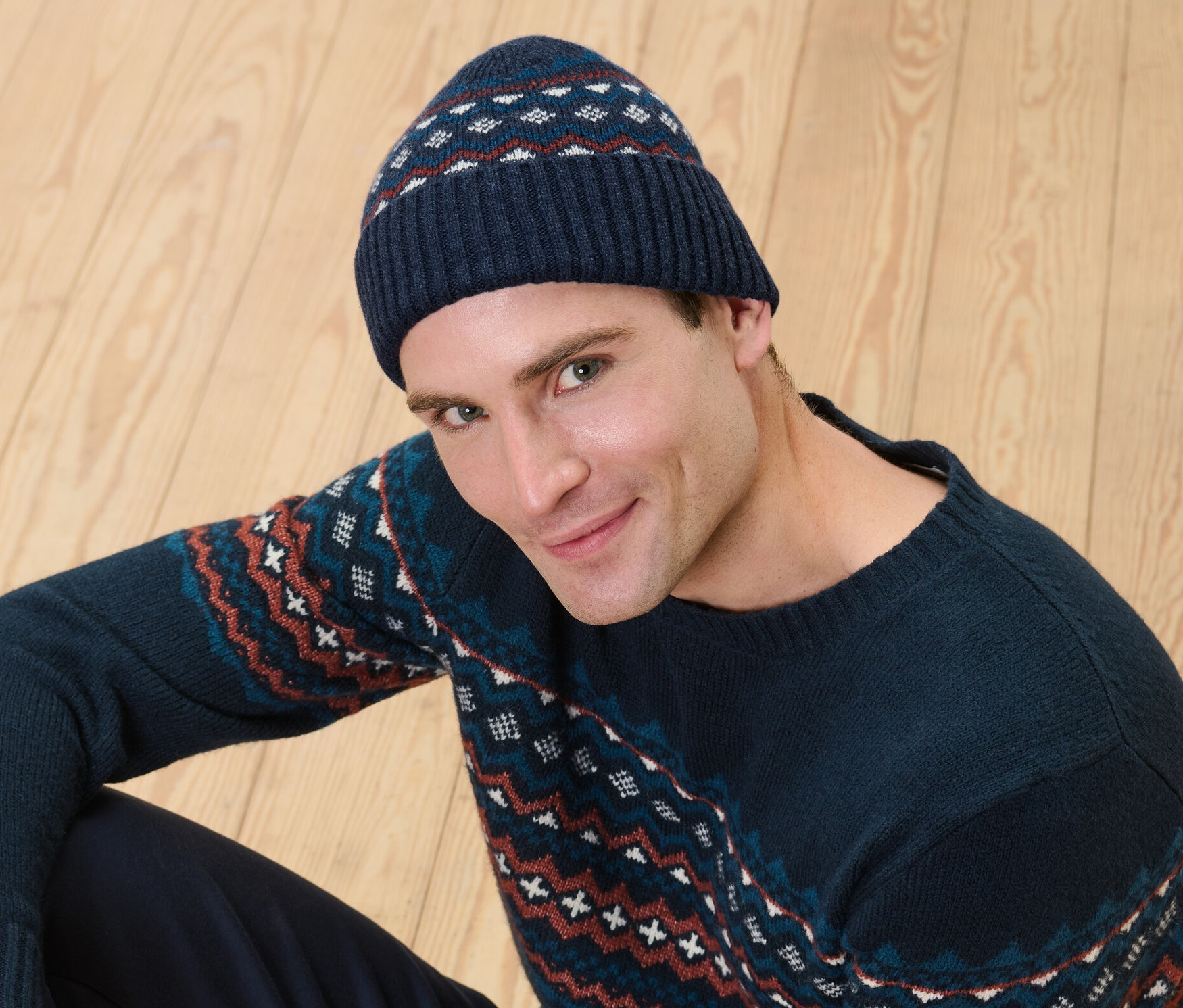 Muž sedí na drevenej podlahe a má na sebe pletenú čiapku s vlnou certifikovanou podľa RWS a sveter so vzorom.