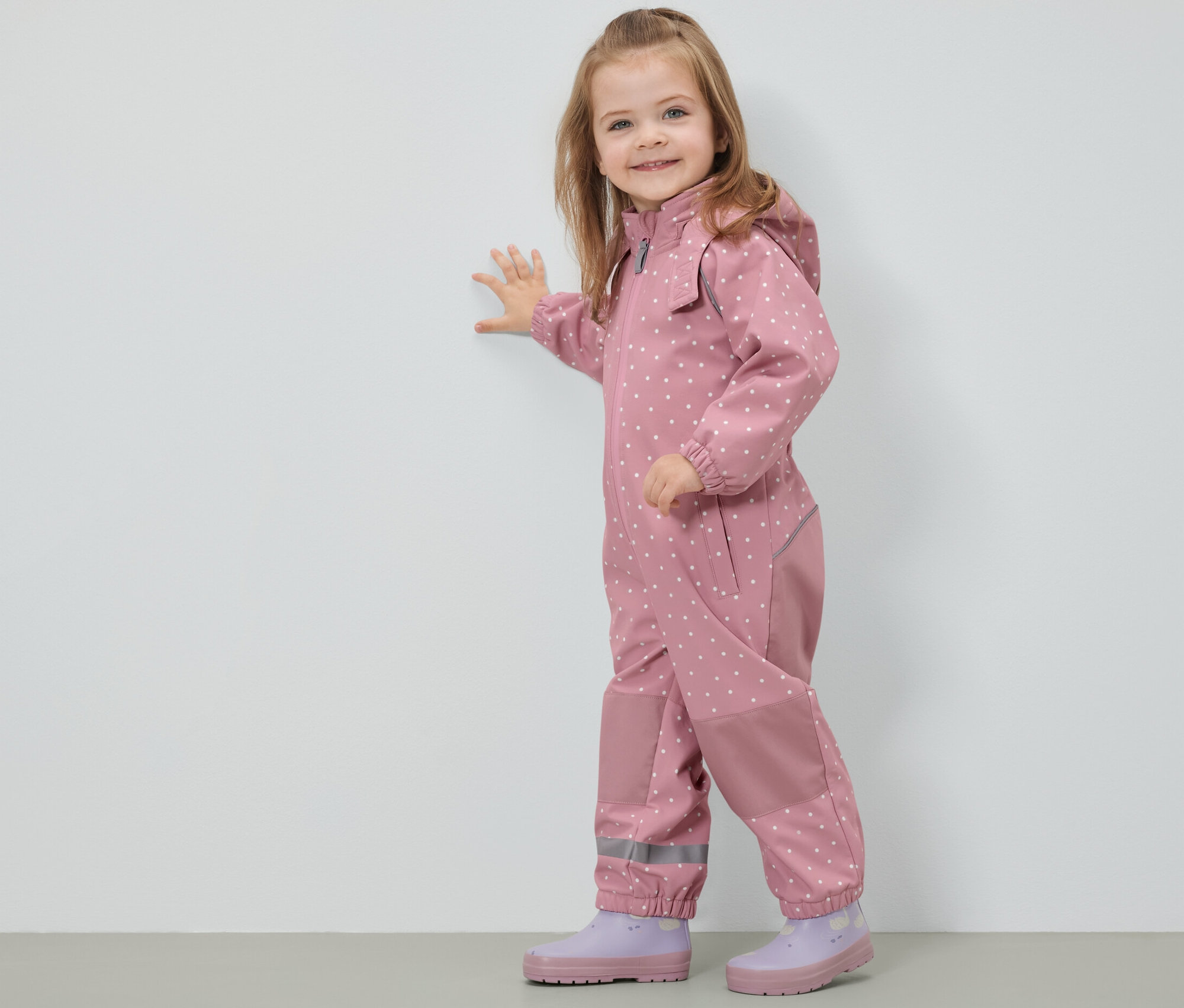 Usmievajúce sa dievčatko v detskej softshellovej kombinéze s bodkami stojí pri stene.