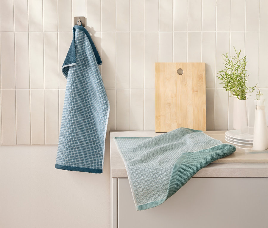 Modrá 2 Waffelpiqué-Küchenhandtücher visí na háku vedľa dosky na krájanie. Ďalšia kuchynská utierka leží na kuchynskej linke.
