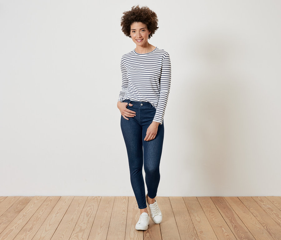 Žena s kučeravými vlasmi pózuje v pruhovanom tričku s dlhým rukávom, tmavomodrých džínsoch a bielych teniskách.
