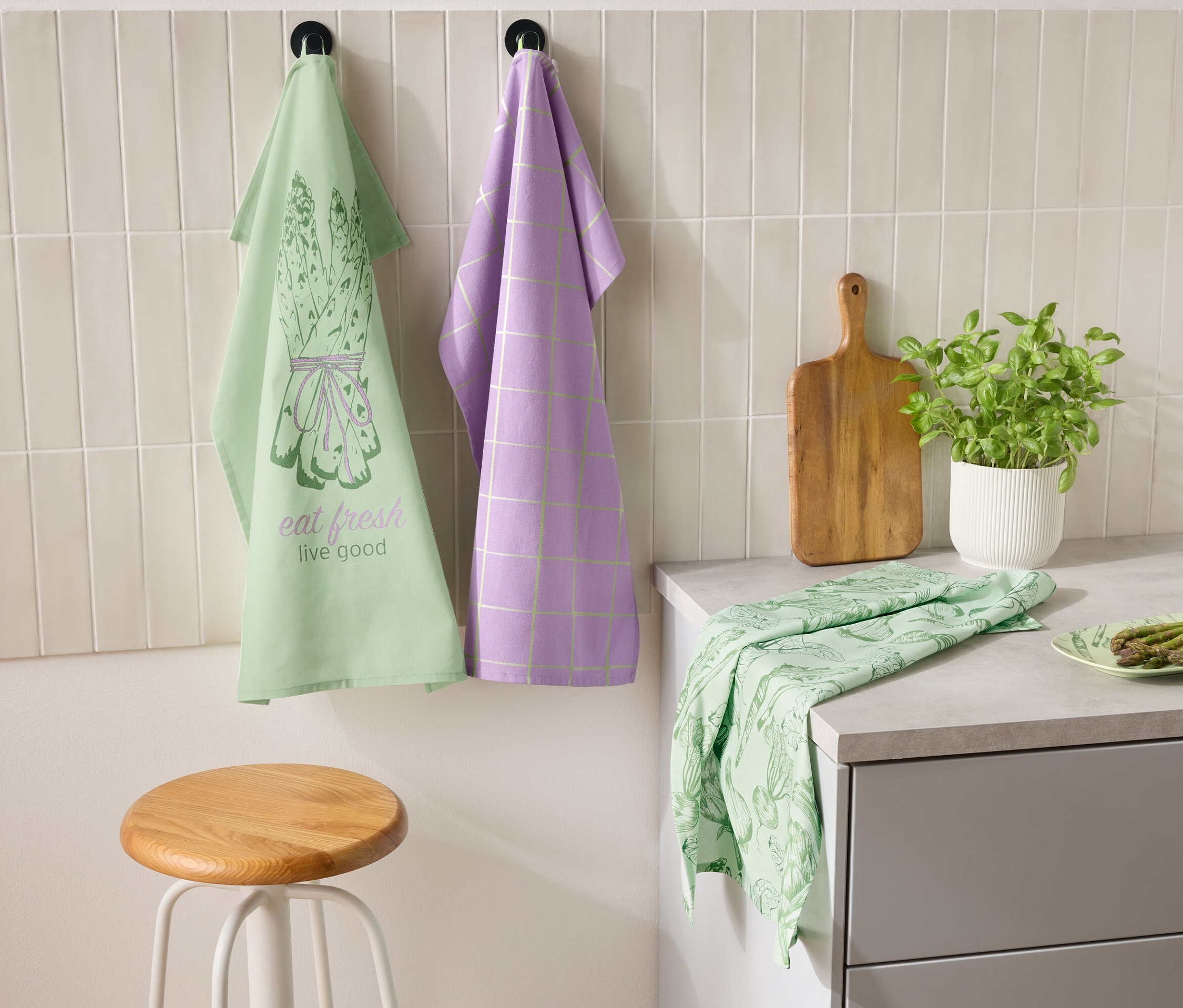 Dve utierky visia vedľa seba na stene, jedna zelená s motívom špargle a nápisom "eat fresh live good" a druhá fialová s kockovaným vzorom. Na linke leží zelená utierka a tanier so špargľou. Vedľa stojí doska na krájanie a kvetináč s bylinkami.