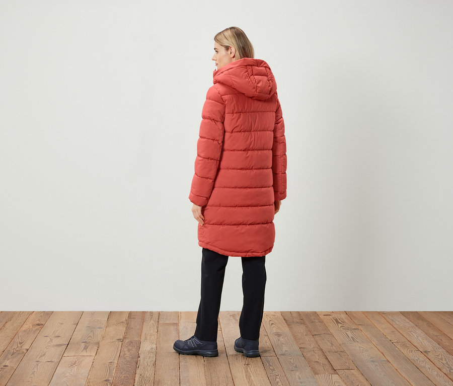 Žena stojí chrbtom, oblečená v červenom prešívanom kabáte s kapucňou, čierne nohavice a tmavé topánky.