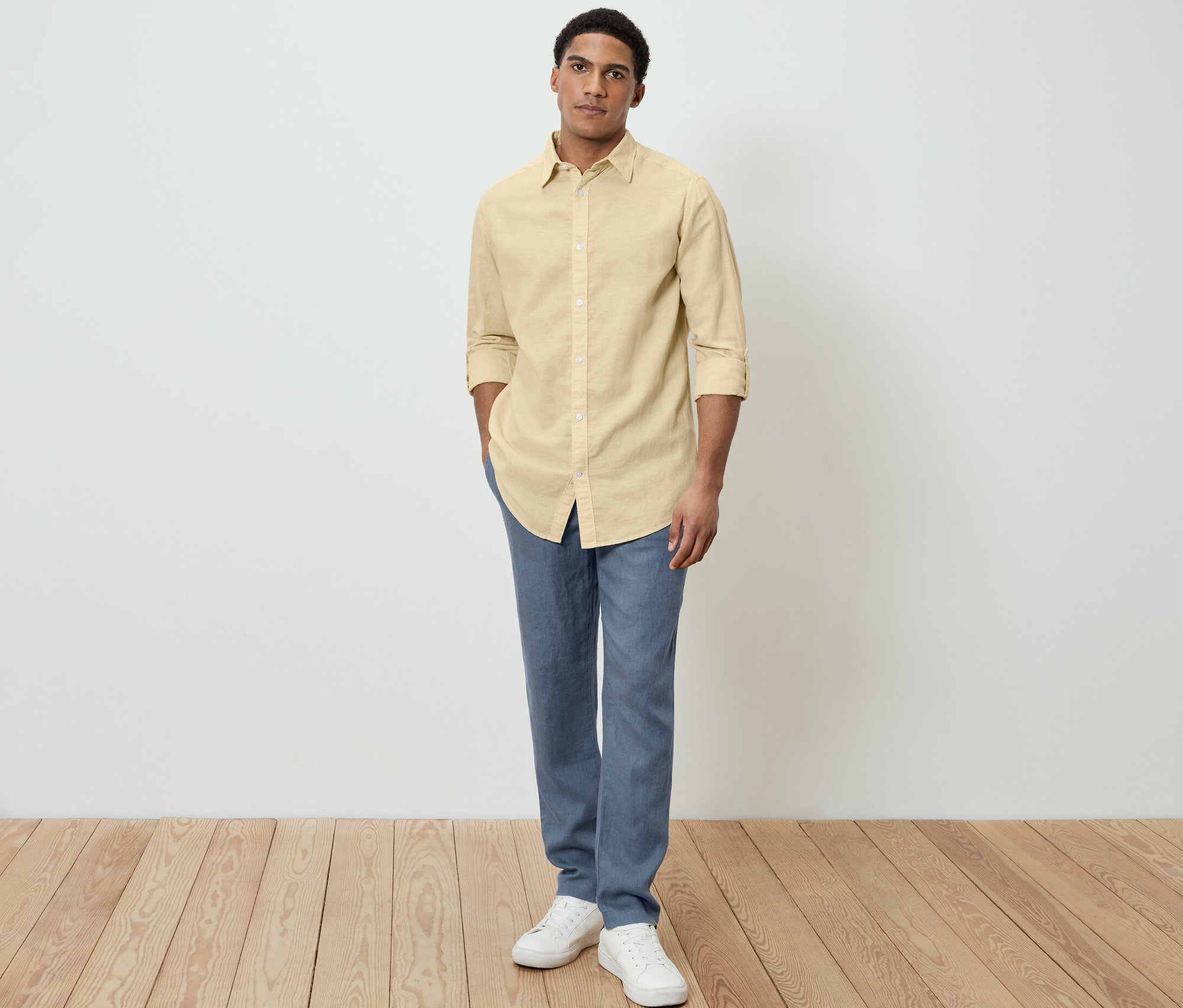 Muž pózuje v žltej košeli s ľanom, modrých nohaviciach a bielych topánkach.