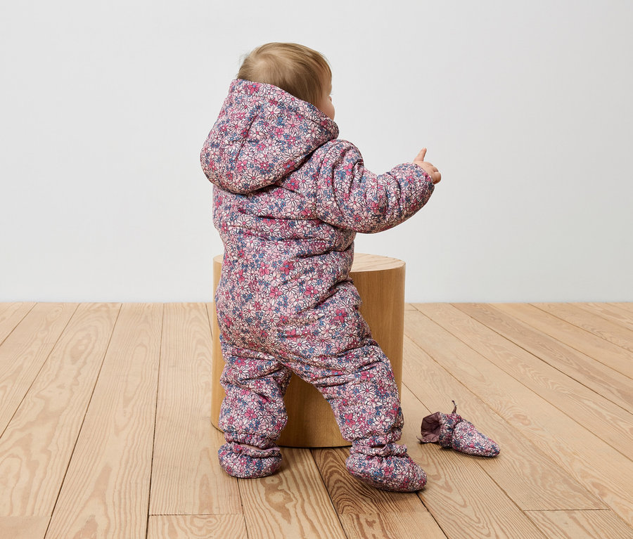 Batoľa sedí na drevenom kláte v kvetinovom overale a ukazuje prstom.