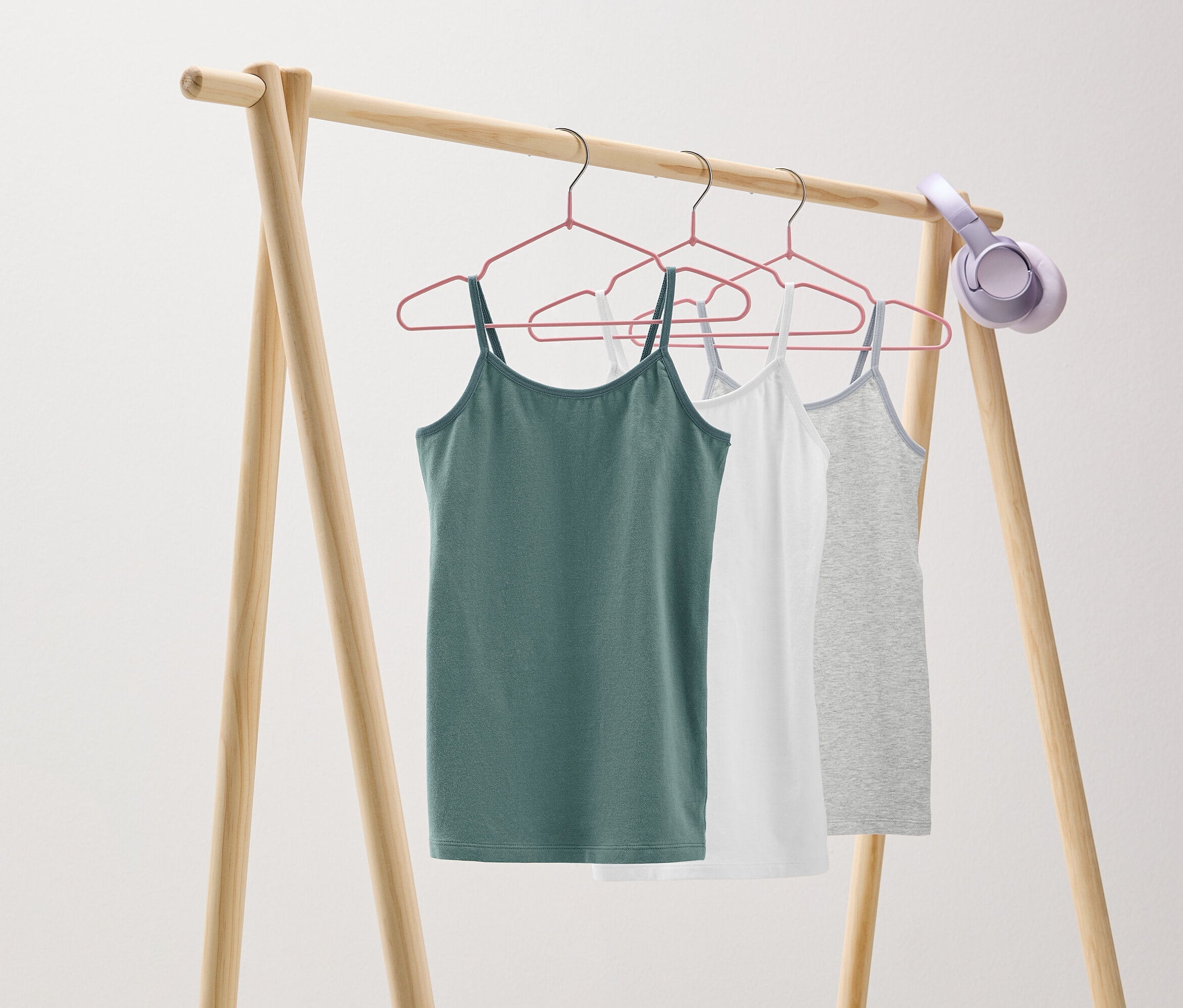 Na drevenom vešiaku visia na ružových vešiakoch tri tielka: zelené, biele a sivé. Na vešiaku sú zavesené slúchadlá.