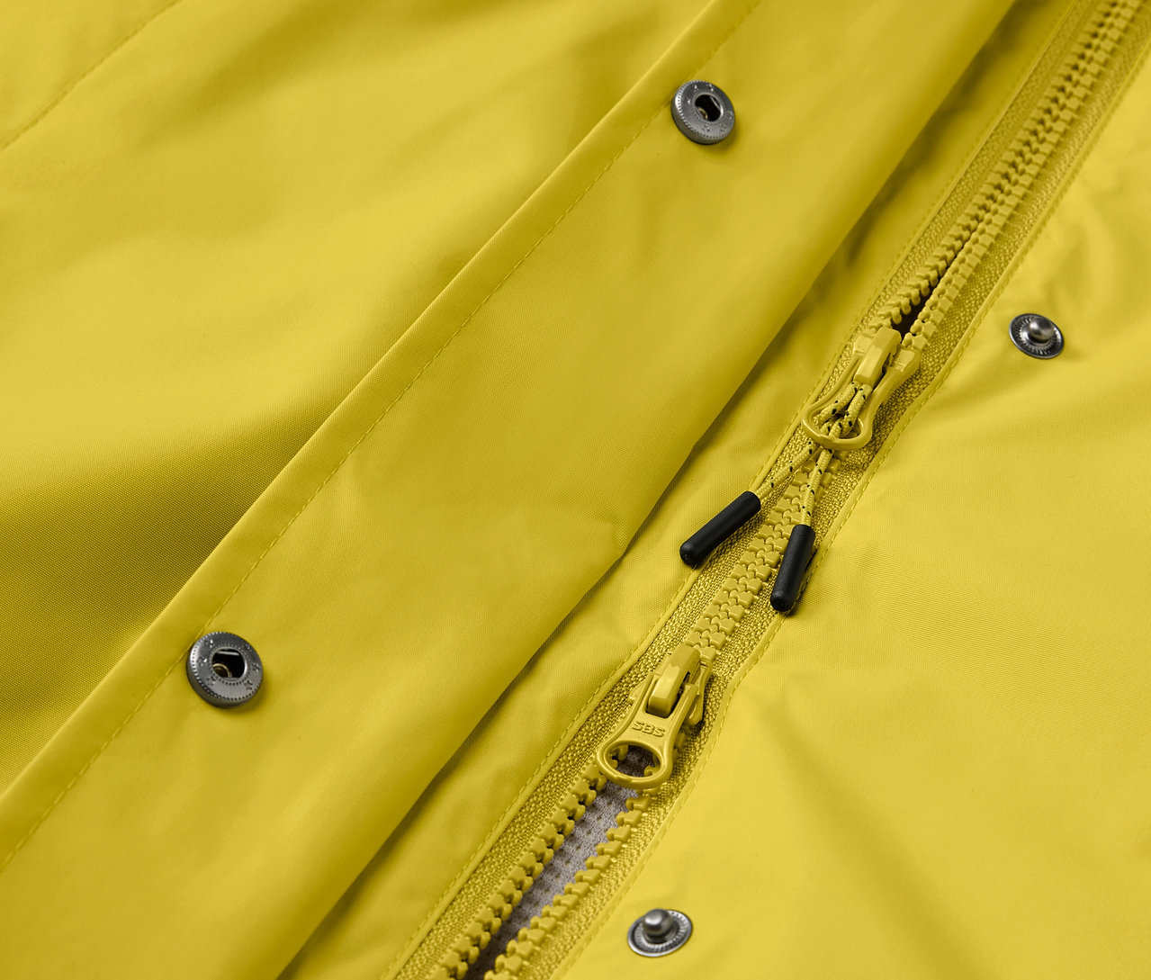 Detail žltej bundy so striebornými patentkami a žltým zipsom.