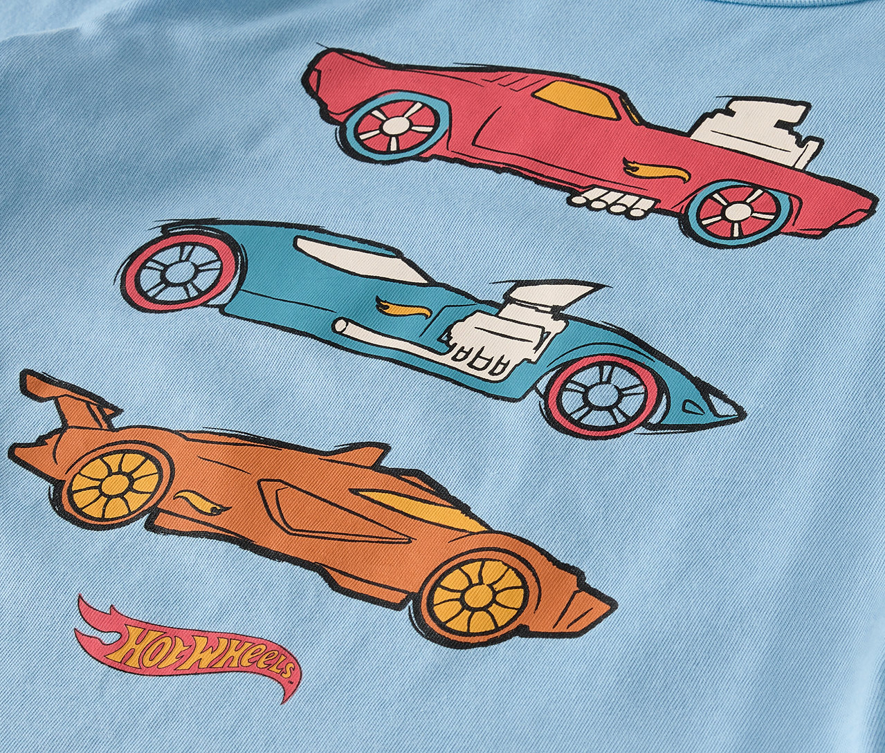 Na svetlomodrom tričku sú tri kreslené autá Hot Wheels v červenej, modrej a oranžovej farbe.