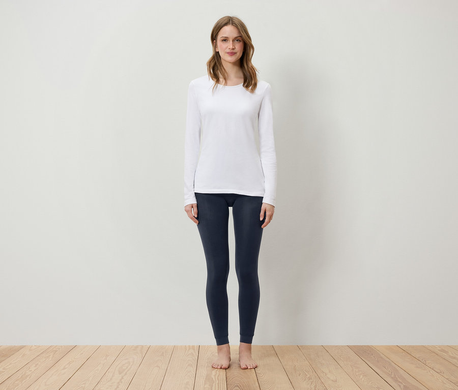 Žena stojí na drevenej podlahe a má na sebe biele tričko s dlhým rukávom a tmavomodré legíny.