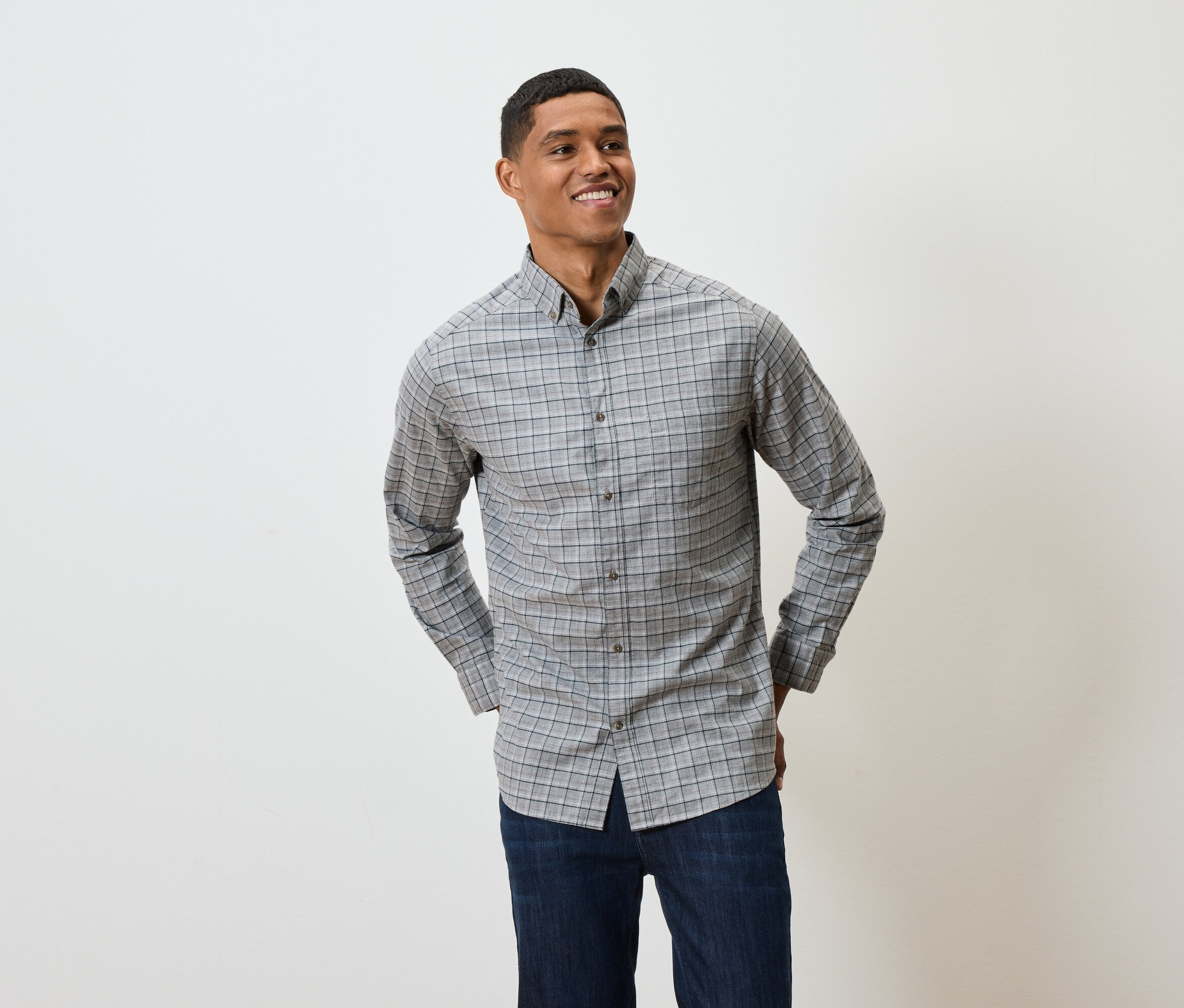 Usmievajúci sa muž pózuje v sivej kockovanej košeli s dlhým rukávom a tmavomodrých džínsoch.