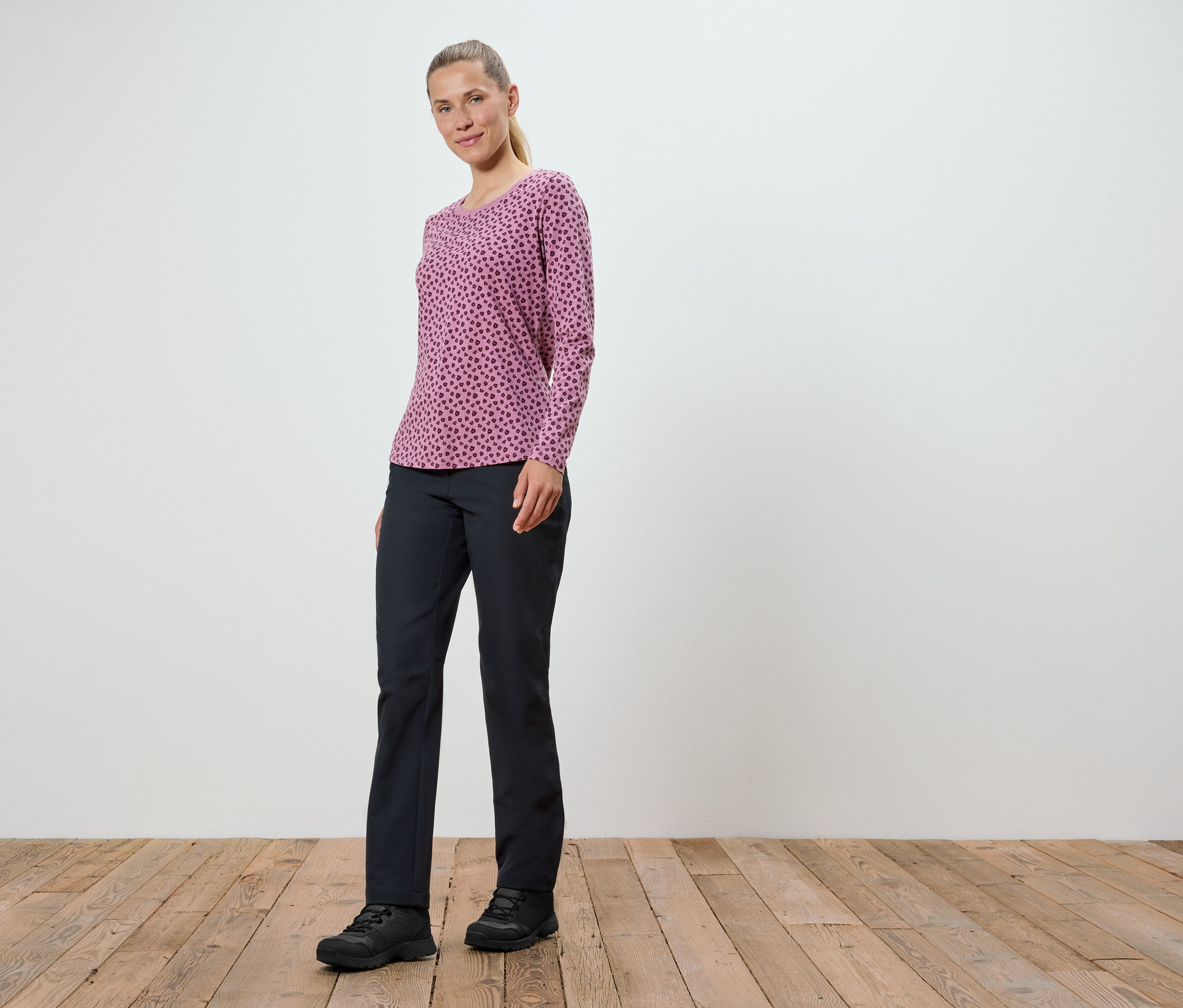 Žena v ružovom tričku s dlhým rukávom a čiernych nohaviciach stojí na drevenej podlahe.