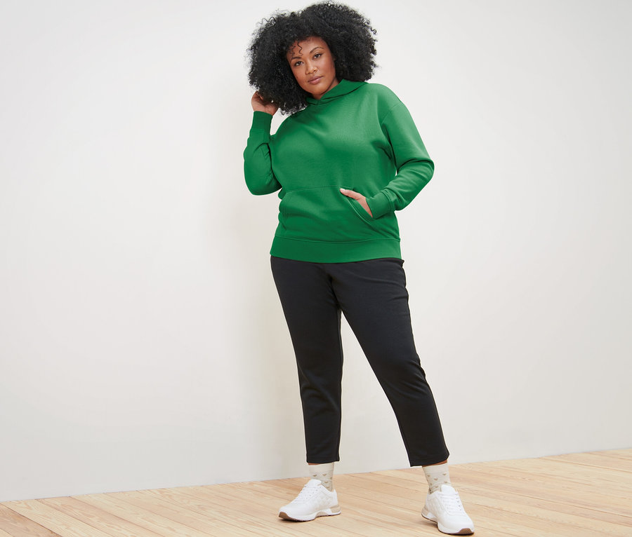 Žena pózuje v zelenej mikine s kapucňou a čiernych nohaviciach.