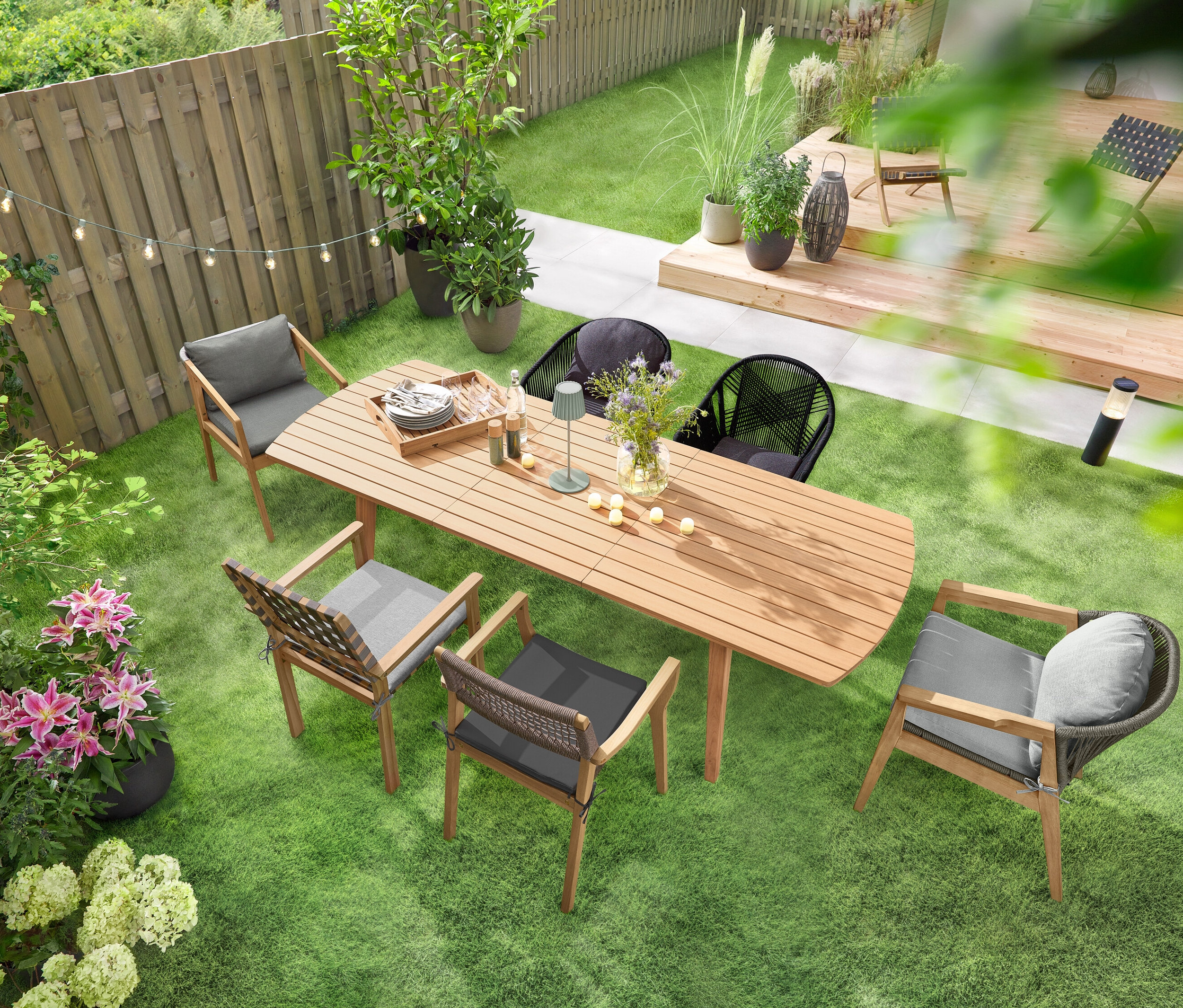 Rozťahovací záhradný stôl so zaoblenými prednými stranami stojí na zelenom trávniku a je obklopený niekoľkými záhradnými stoličkami. Nad stolom visí LED svetelná reťaz »Glühbirnen« a na ňom stojí LED akumulátorová stolová lampa do exteriéru, šalviová. Na stole je rozmiestnených niekoľko LED akumulátorových čajových sviečok do exteriéru, 6 ks.