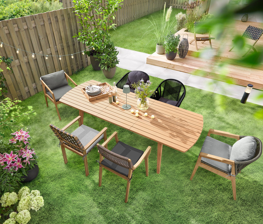 Rozťahovací záhradný stôl so zaoblenými prednými stranami stojí na zelenom trávniku a je obklopený niekoľkými záhradnými stoličkami. Nad stolom visí LED svetelná reťaz »Glühbirnen« a na ňom stojí LED akumulátorová stolová lampa do exteriéru, šalviová. Na stole je rozmiestnených niekoľko LED akumulátorových čajových sviečok do exteriéru, 6 ks.