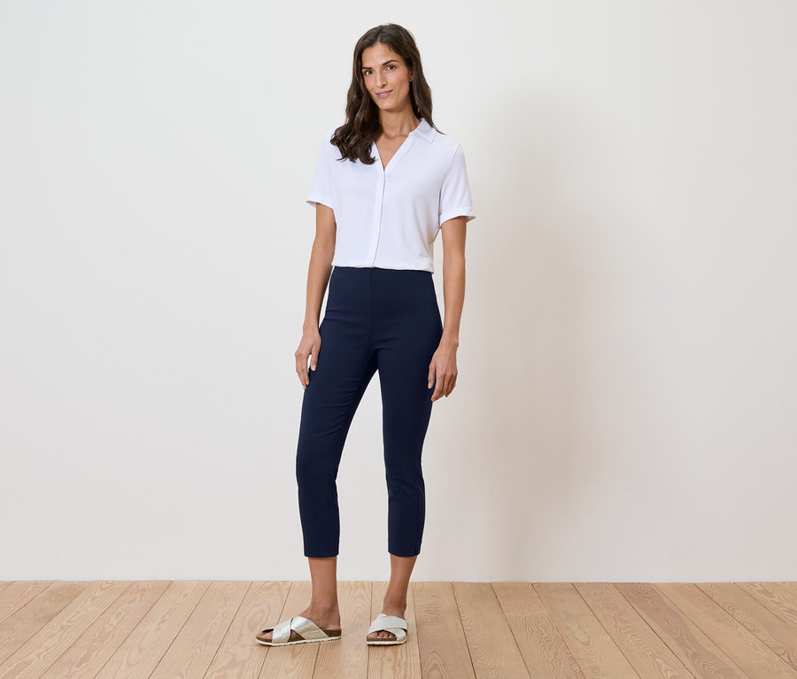 Žena stojí pred stenou a má na sebe bielu košeľu, modré nohavice a zlaté sandále.