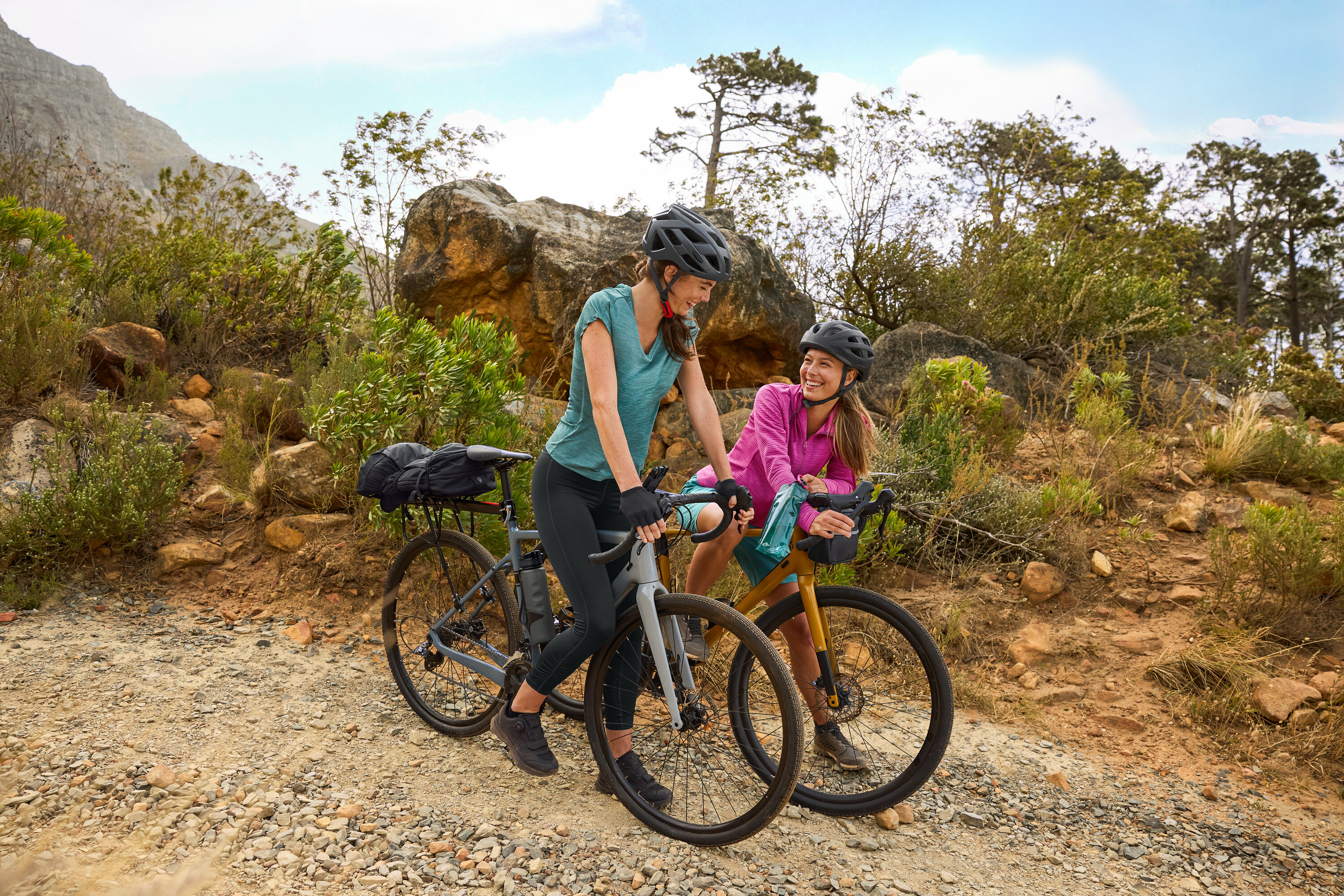 Dve ženy s prilbami stoja s bicyklami na štrkovej ceste v kopcovitom teréne. Na bicykli je pripevnená fľaša na pitie.