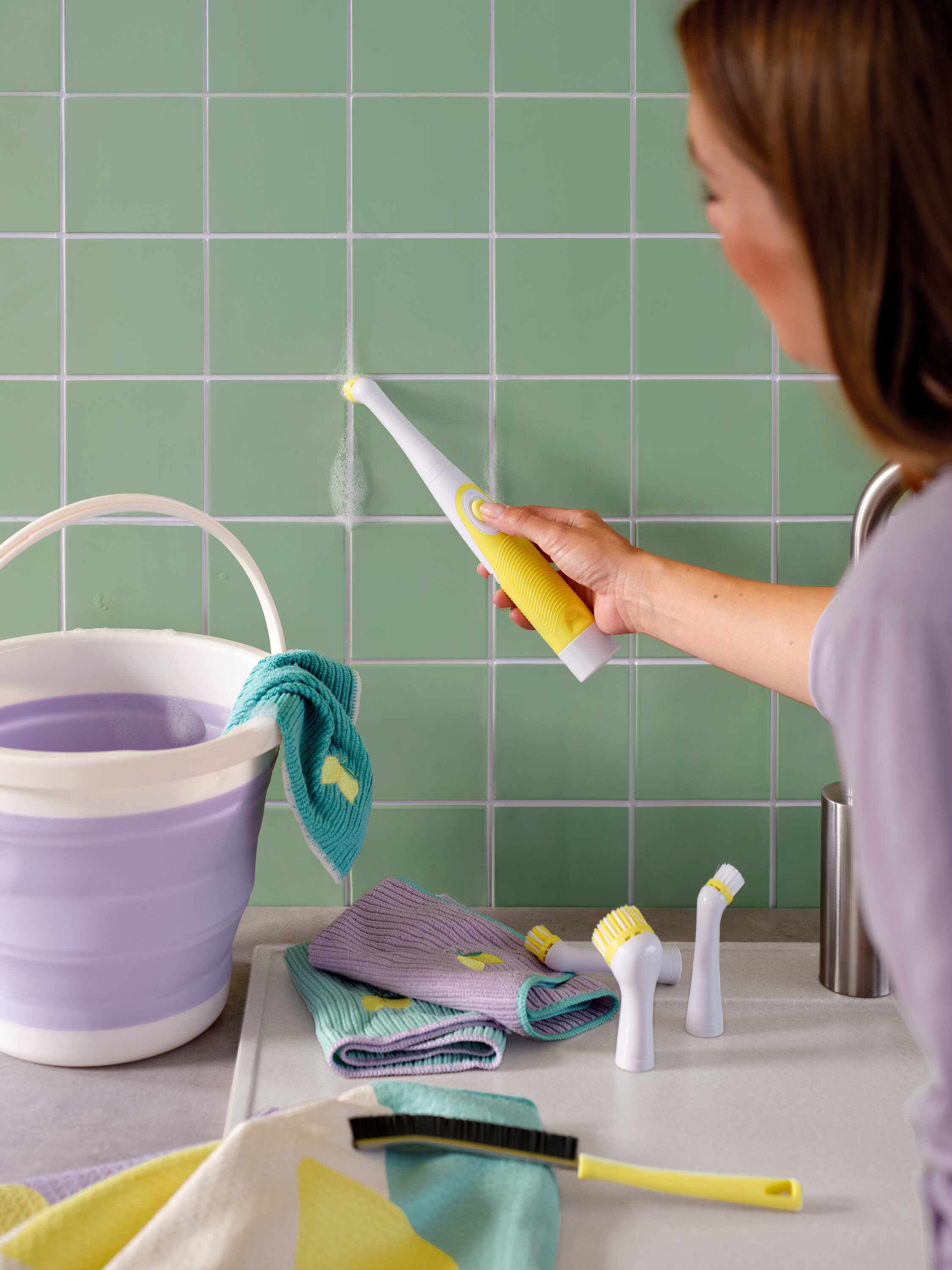 Žena čistí zelené dlaždice žlto-bielou kefou, vidieť aj vedro a handry.