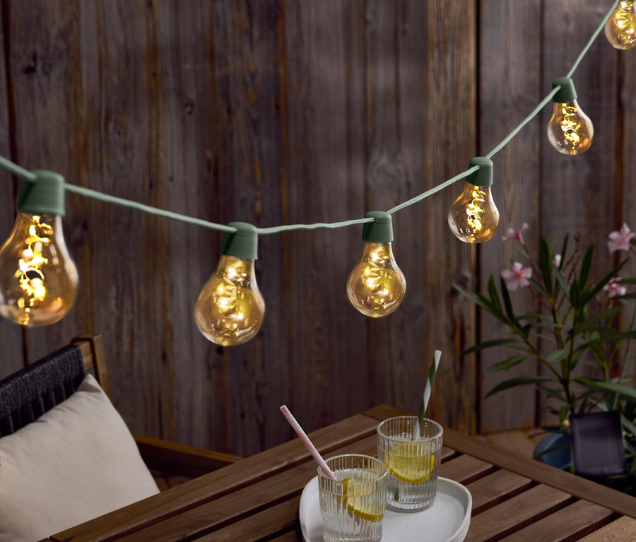 Svetelná reťaz so žiarovkami visí nad dreveným stolom s nápojmi a klobúkom.