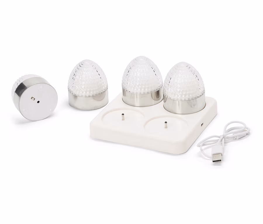 Štyri malé okrúhle lampy so striebornými základňami a fasetovanými, priehľadnými sklenenými kupolami stoja na bielej nabíjacej stanici s USB káblom.