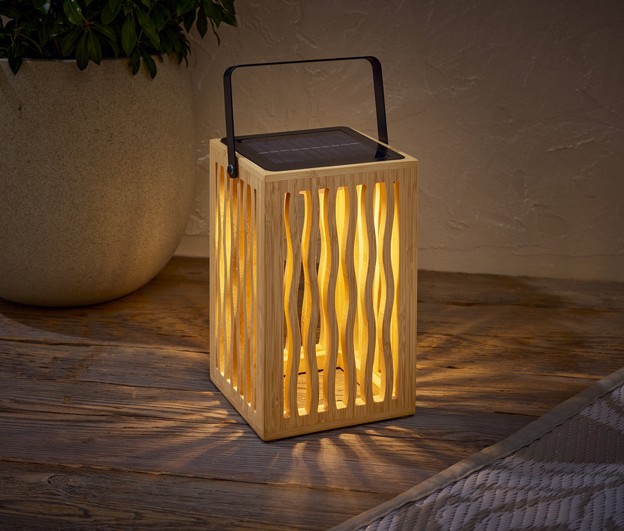 Drevená solárna lampa s čiernou rúčkou stojí na drevenom stole vedľa kvetináča s rastlinou.