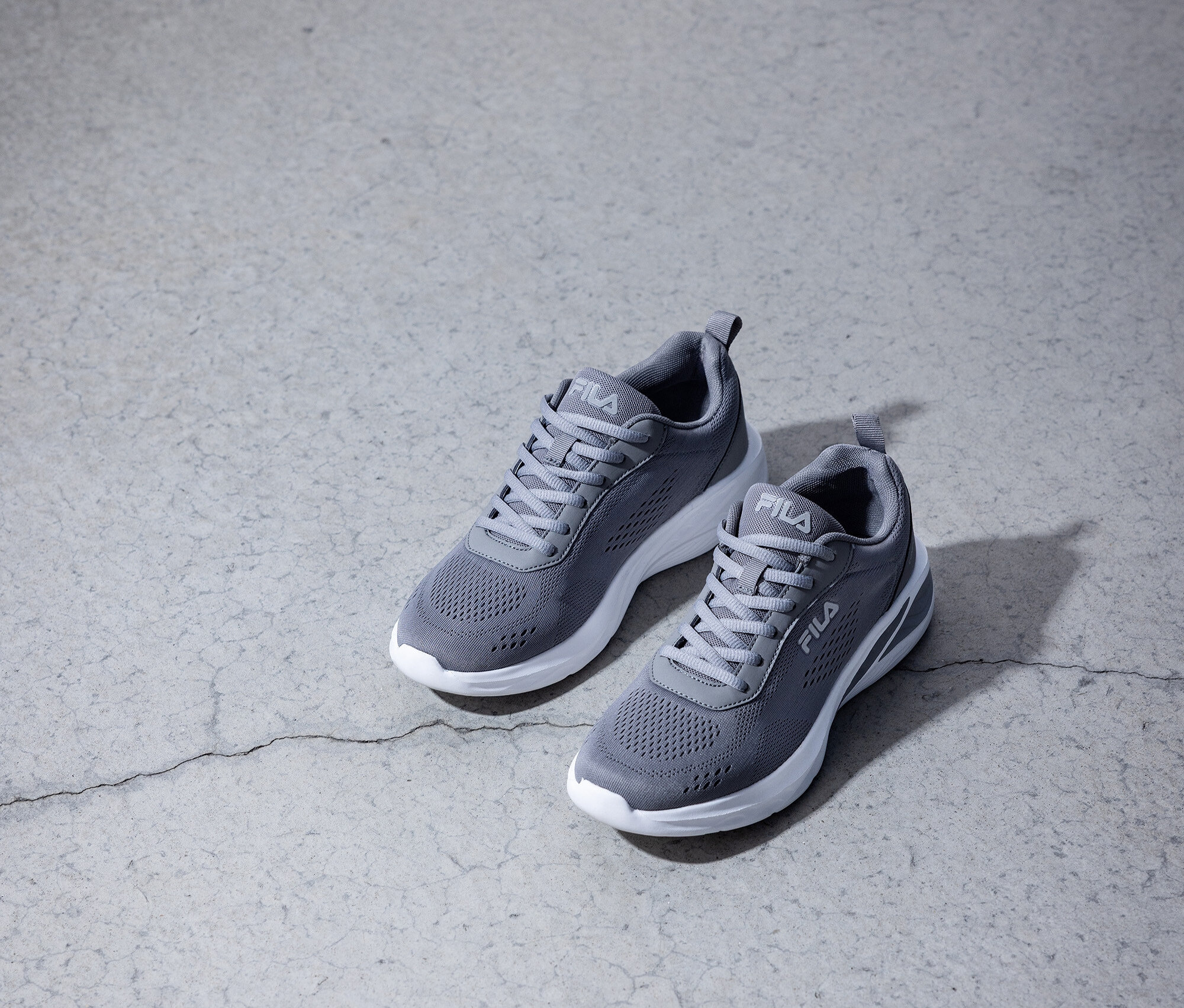 Dva sivé tenisky Fila s bielymi podrážkami stoja na sivej betónovej podlahe.