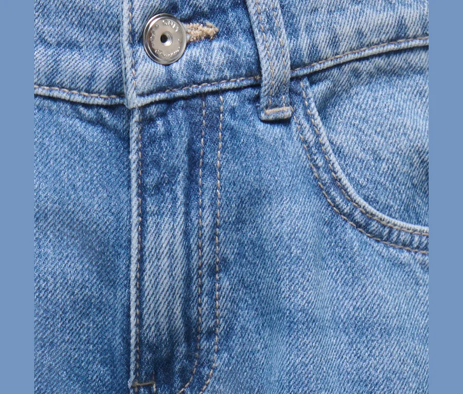 Detail modrých džínsov zobrazujúci strieborný gombík, gombíkovú dierku, rázporok, pútko na opasok a predné vrecko s béžovým prešívaním.
