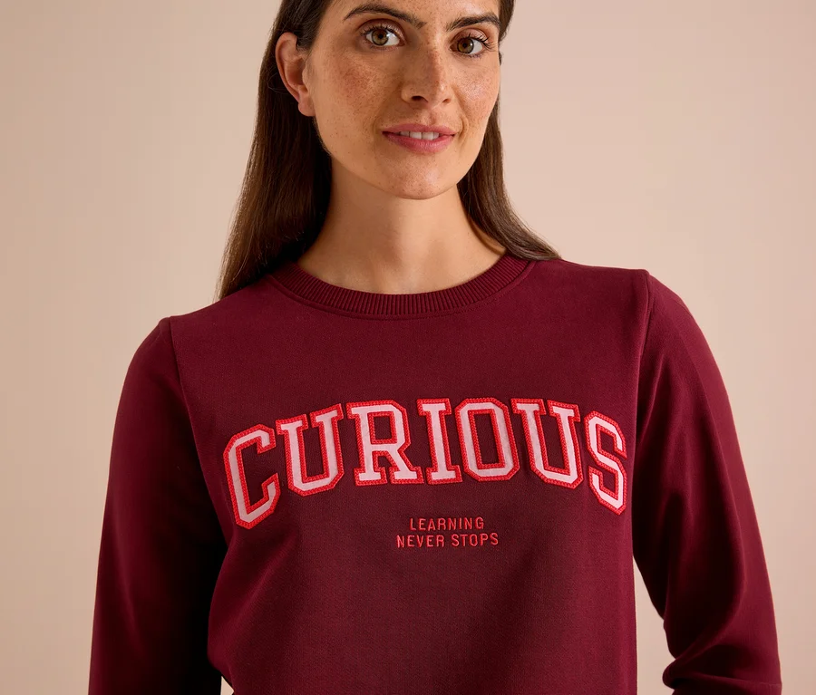 Žena má na sebe bordovú mikinu CECIL s nápisom „CURIOUS“ a „LEARNING NEVER STOPS“.