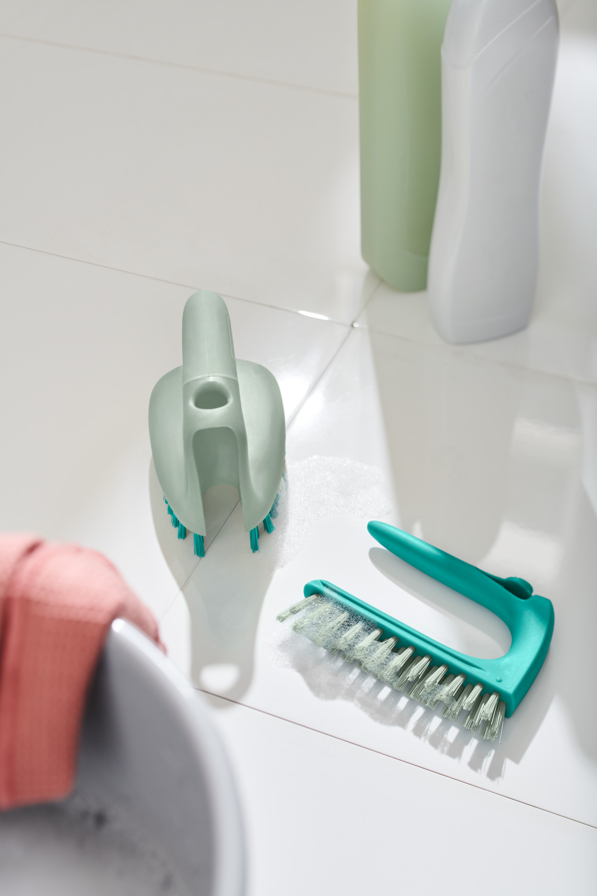 Dve kefy na čistenie obkladačiek a podláh 2 v 1 ležia na bielych dlaždiciach vedľa umývadla.