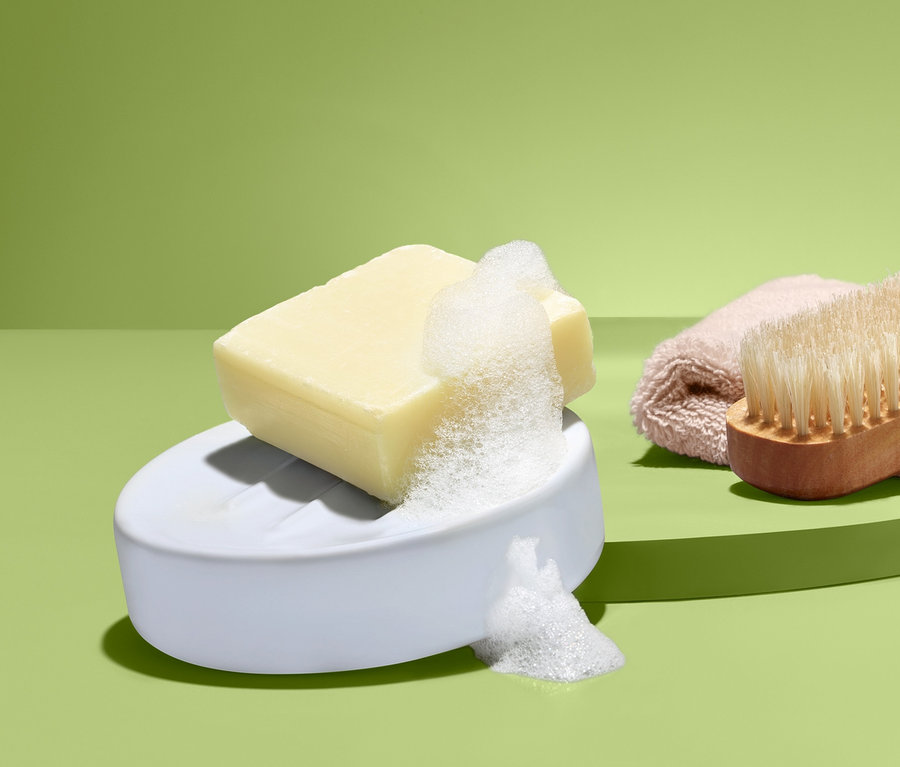Na zelenom pozadí je mydlo s penou na bielej miske na mydlo, vedľa kefa a uterák.