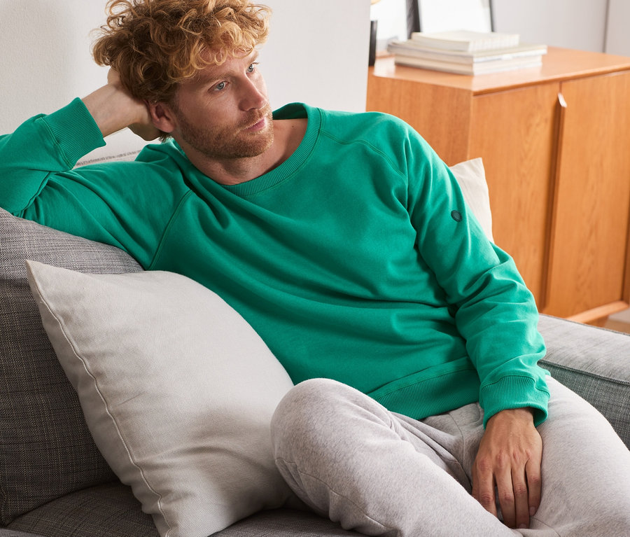 Muž leží na sivej pohovke a má na sebe zelenú mikinu.