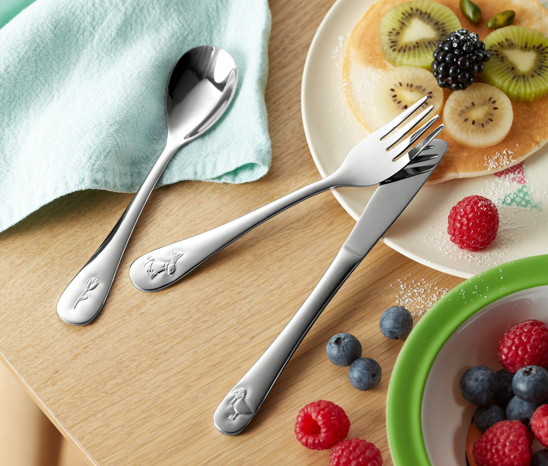 Na drevenom stole leží detský príbor (3-dielna súprava), tanier s palacinkami a ovocím, misa s čučoriedkami a malinami a pohár s nápojom.