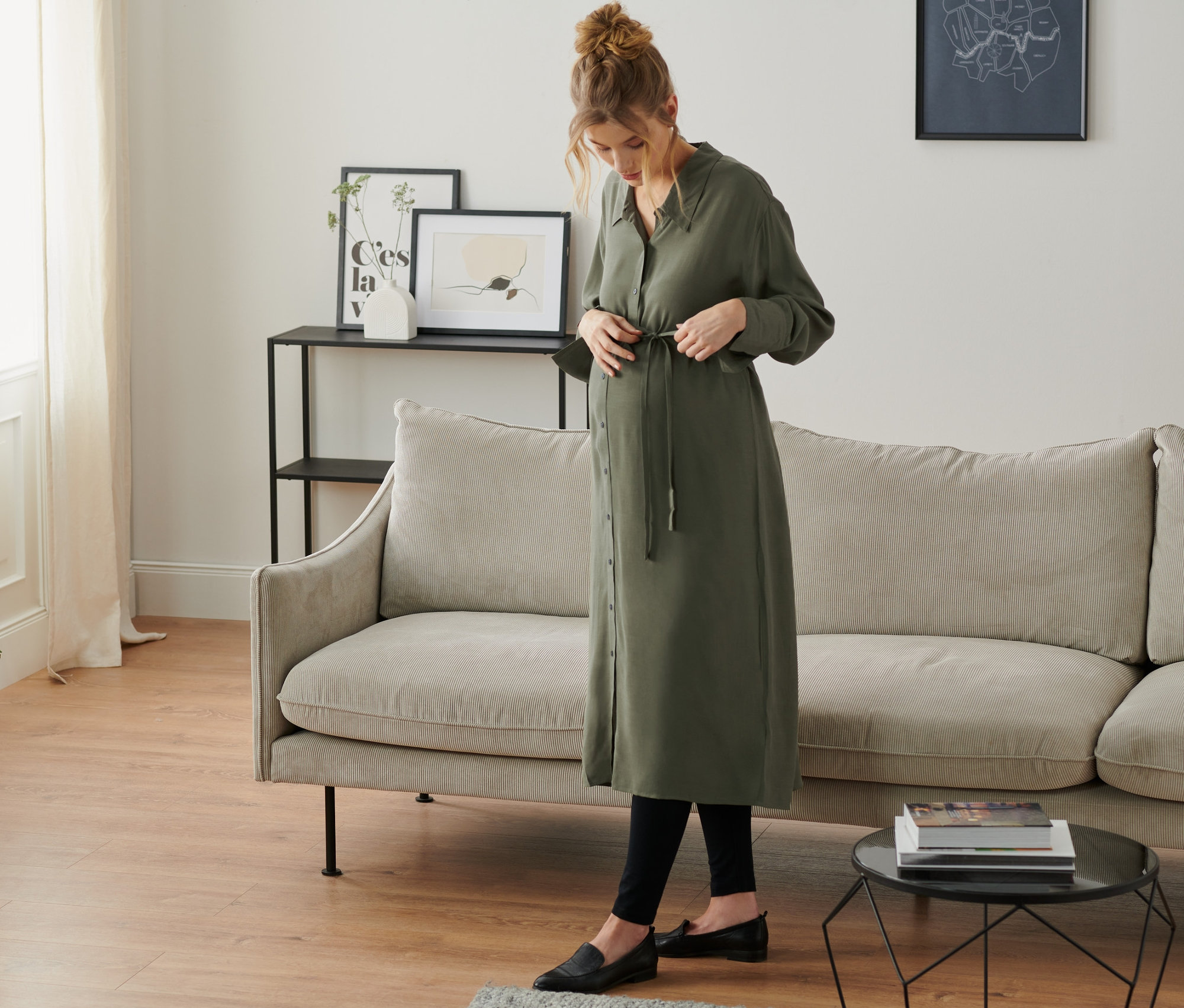Tehotná žena si zaväzuje olivové košeľové šaty v obývacej izbe.