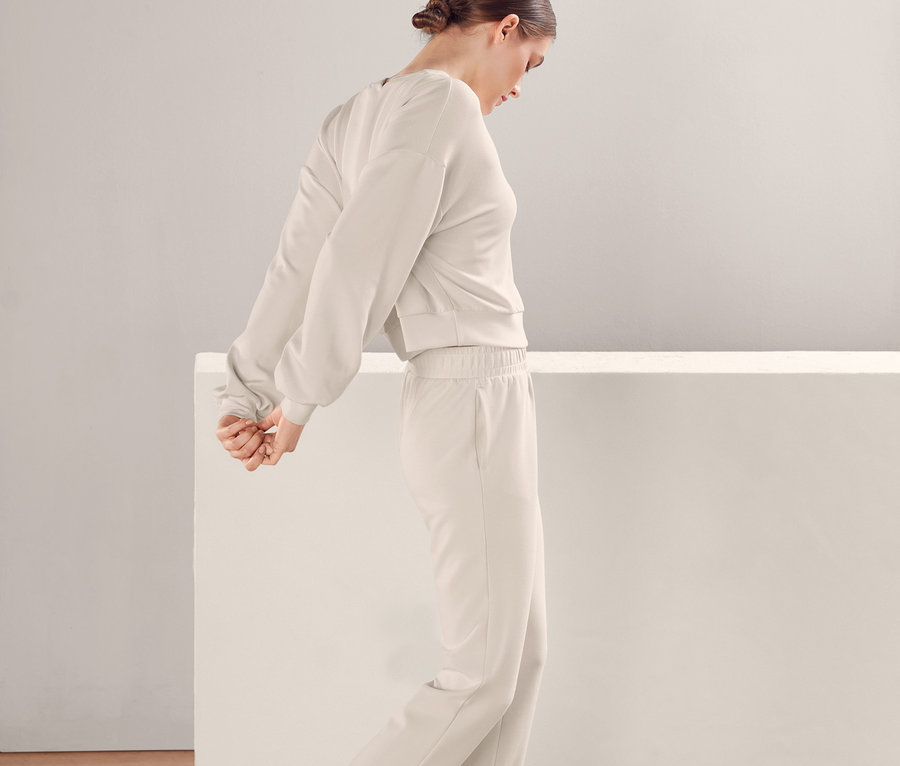 Žena v béžovej teplákovej súprave a teniskách stojí pred bielym múrom. Má na sebe Obzvlášť mäkkú mikinu.