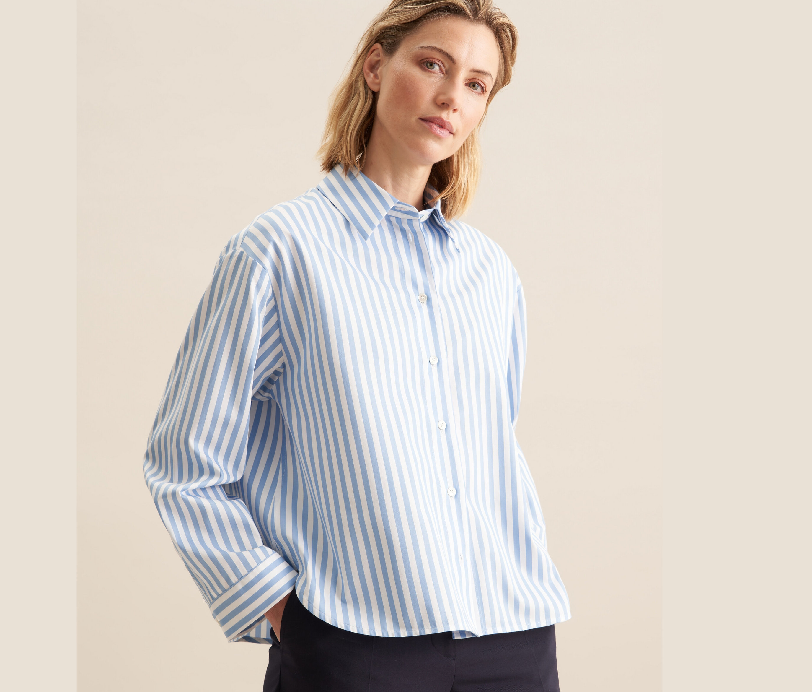Žena má na sebe modrú košeľovú blúzku nadmernej veľkosti Seidensticker s bielymi prúžkami a tmavomodré nohavice.