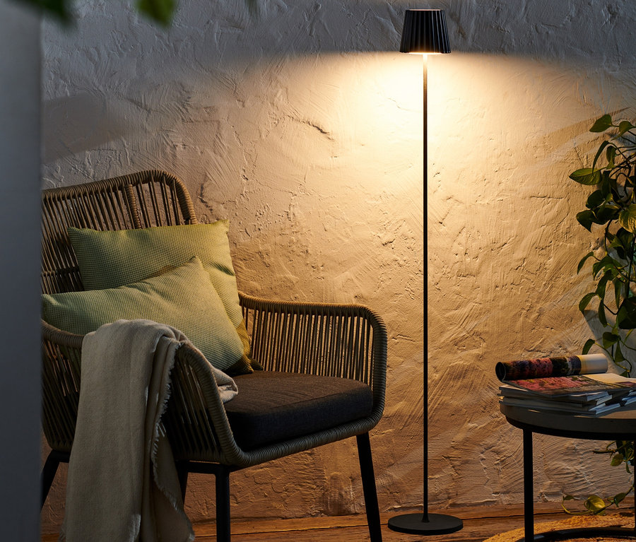 Čierna stojacia lampa LED na akumulátor stojí vedľa prúteného kresla s vankúšmi.