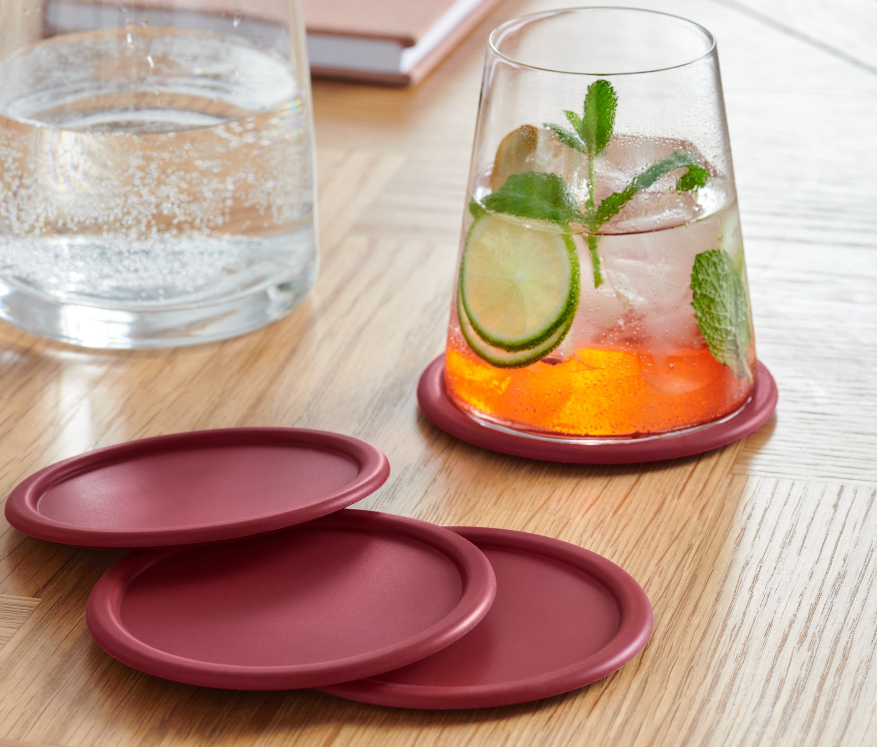 Pohár s nápojom stojí na jednom zo štyroch bordových silikónových podložiek. Ďalšie tri podložky ležia vedľa seba na drevenom stole. V pozadí je karafa s vodou a notes.