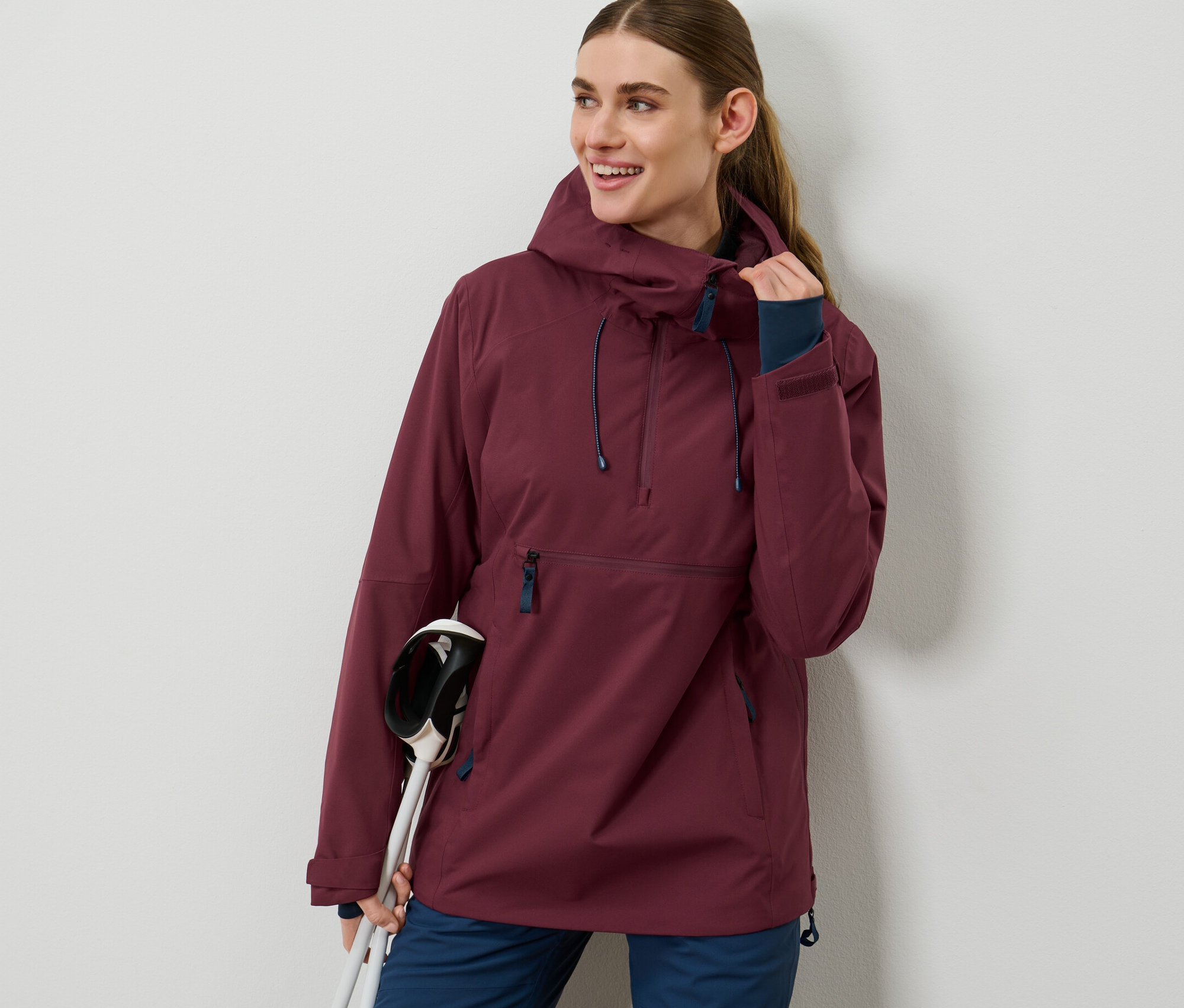 Žena má na sebe bordovú lyžiarsku bundu s polovičným zipsom a drží lyžiarske palice.