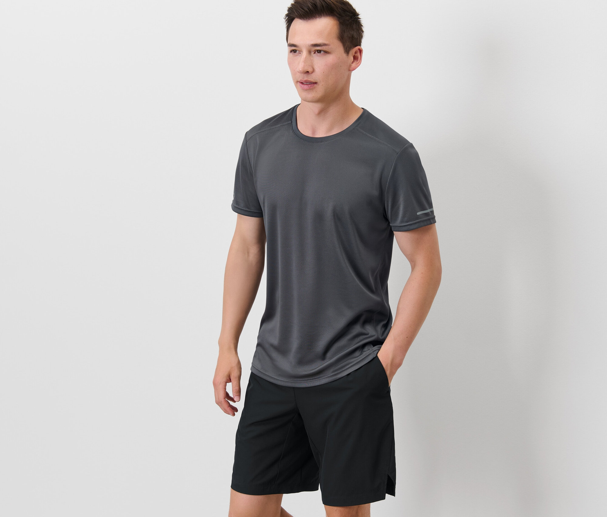 Muž stojí a má ruky vo vreckách čiernych šortiek, na sebe má sivé funkčné tričko.