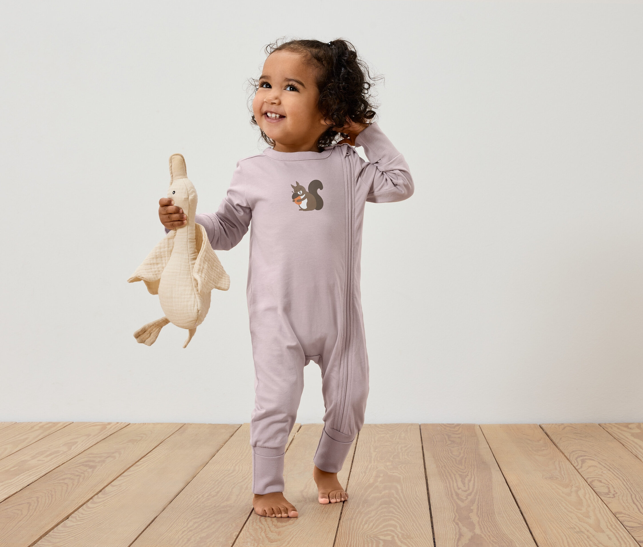 Dieťa v ružovom overale so zipsom a obrázkom veveričky drží plyšovú hračku husi.